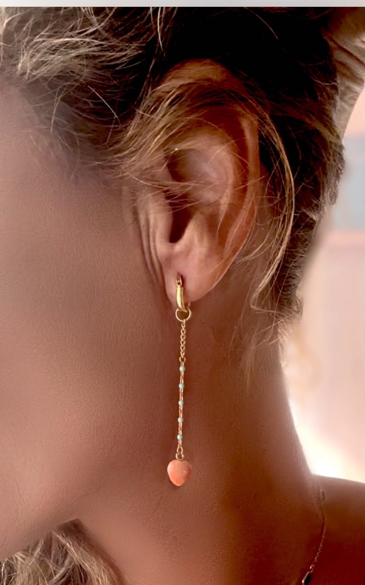 Woman wearing a delicate gold mini hoop dangle earring with a love heart shape attached at the bottom in angel skin coral with enamel beads along gold chain.