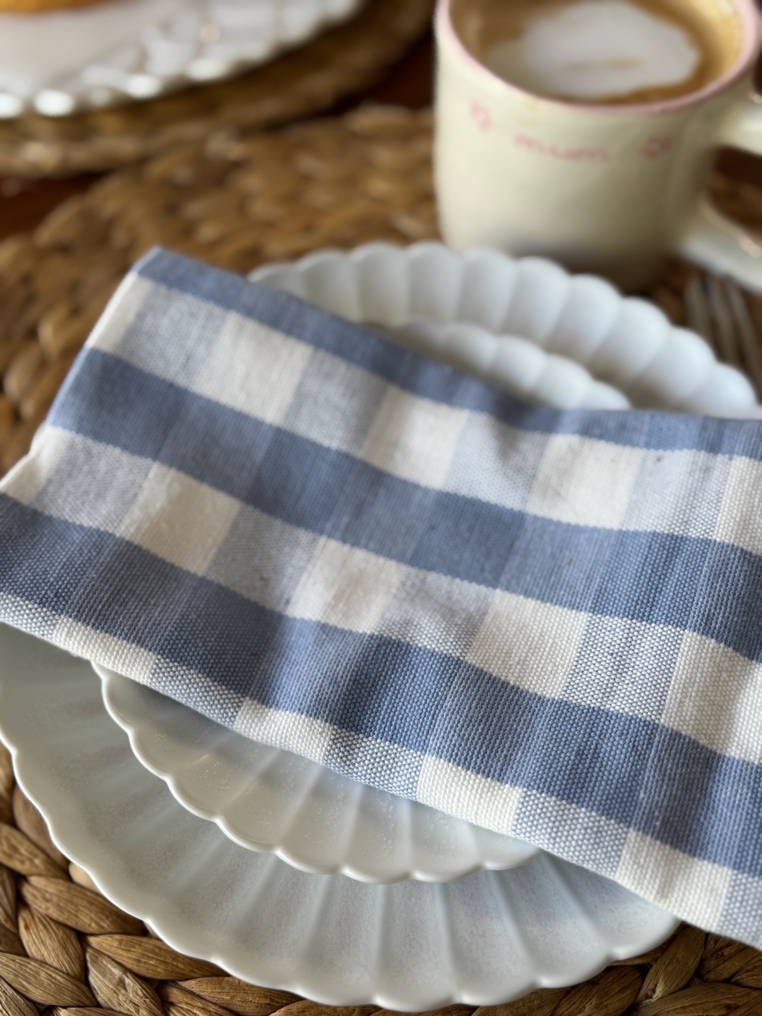 Blue and white checkered cloth napkin on a white plate with a cup of coffee in the background.