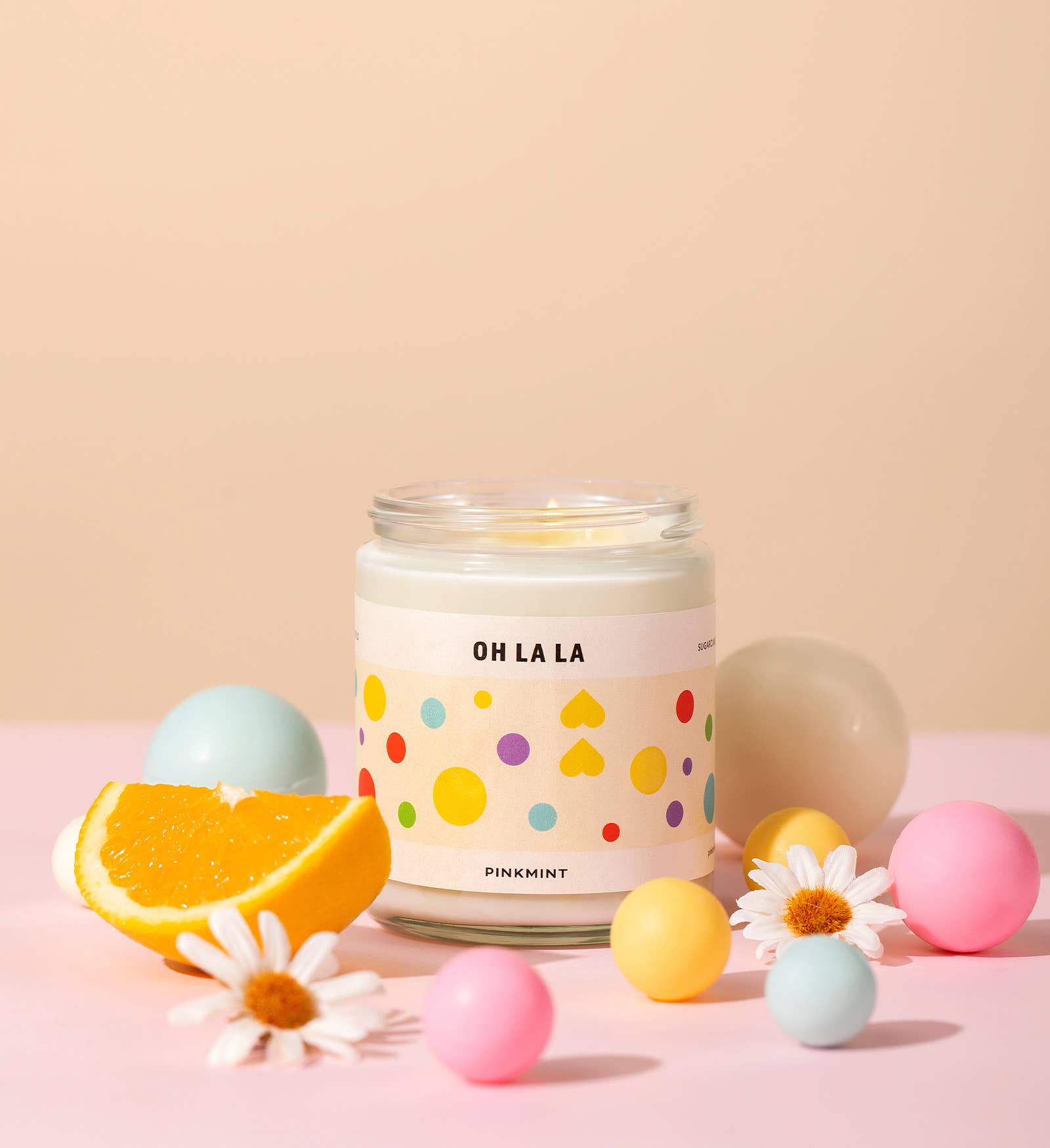 White soy candle in a jar with a wick lit and an orange sliced and flowers around it on a table - ANNIE.G