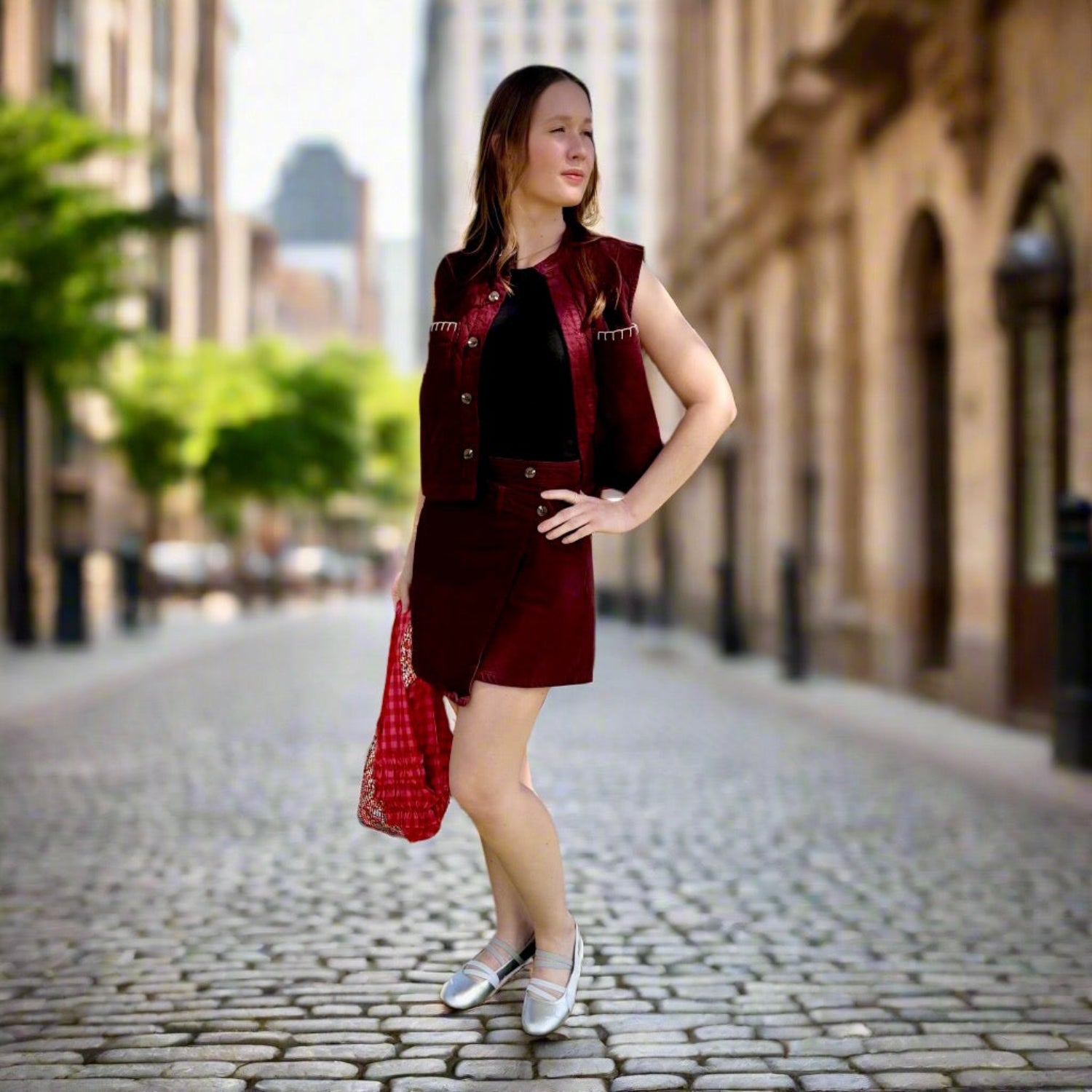 Woman in a mulberry mini skirt and quilted vest standing on a cobblestone street carrying a tote with blurred background