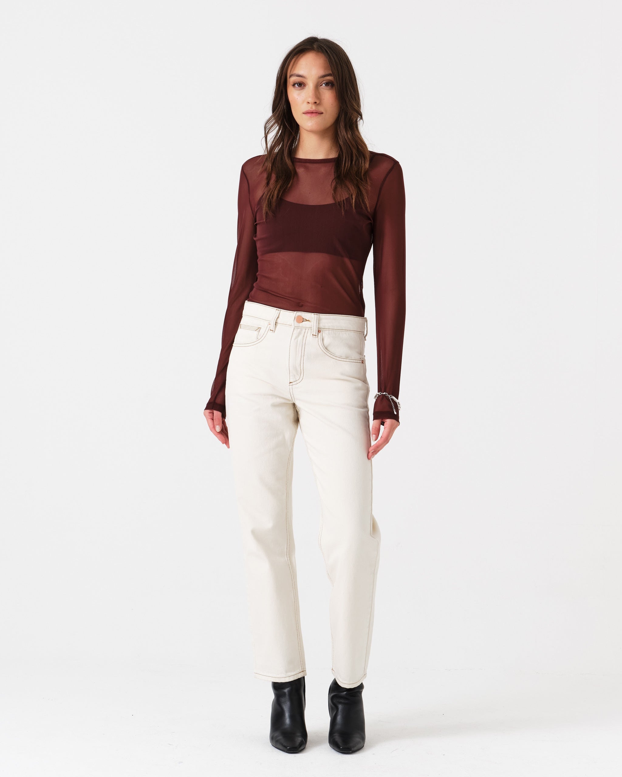 Woman wearing a burgundy sheer top and white jean trousers with contrast stitching, pockets, with a zip and button closure and belt loops.