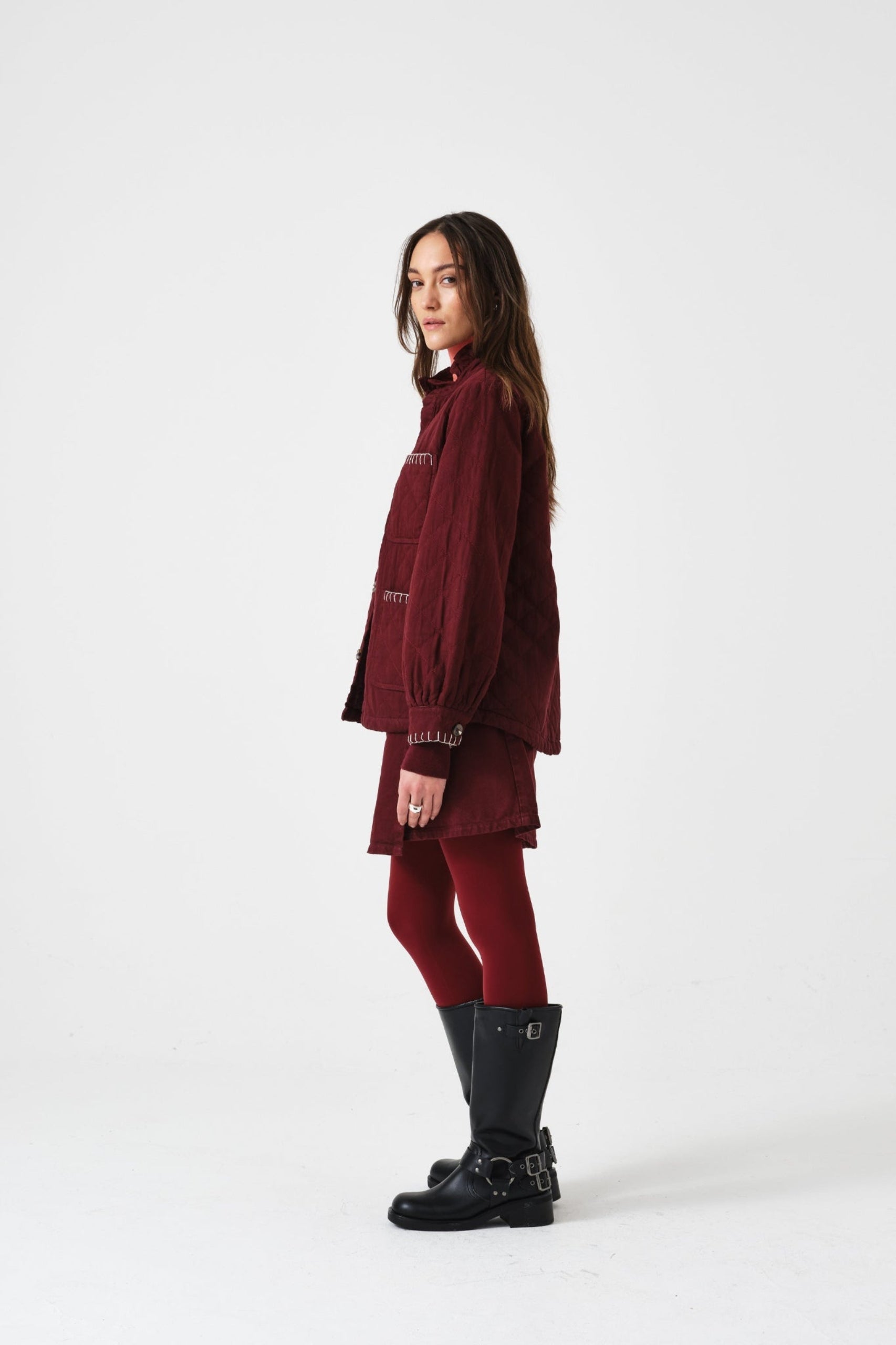 Person standing on the side wearing a burgundy mini skirt and quilted jacket outfit with black boots.