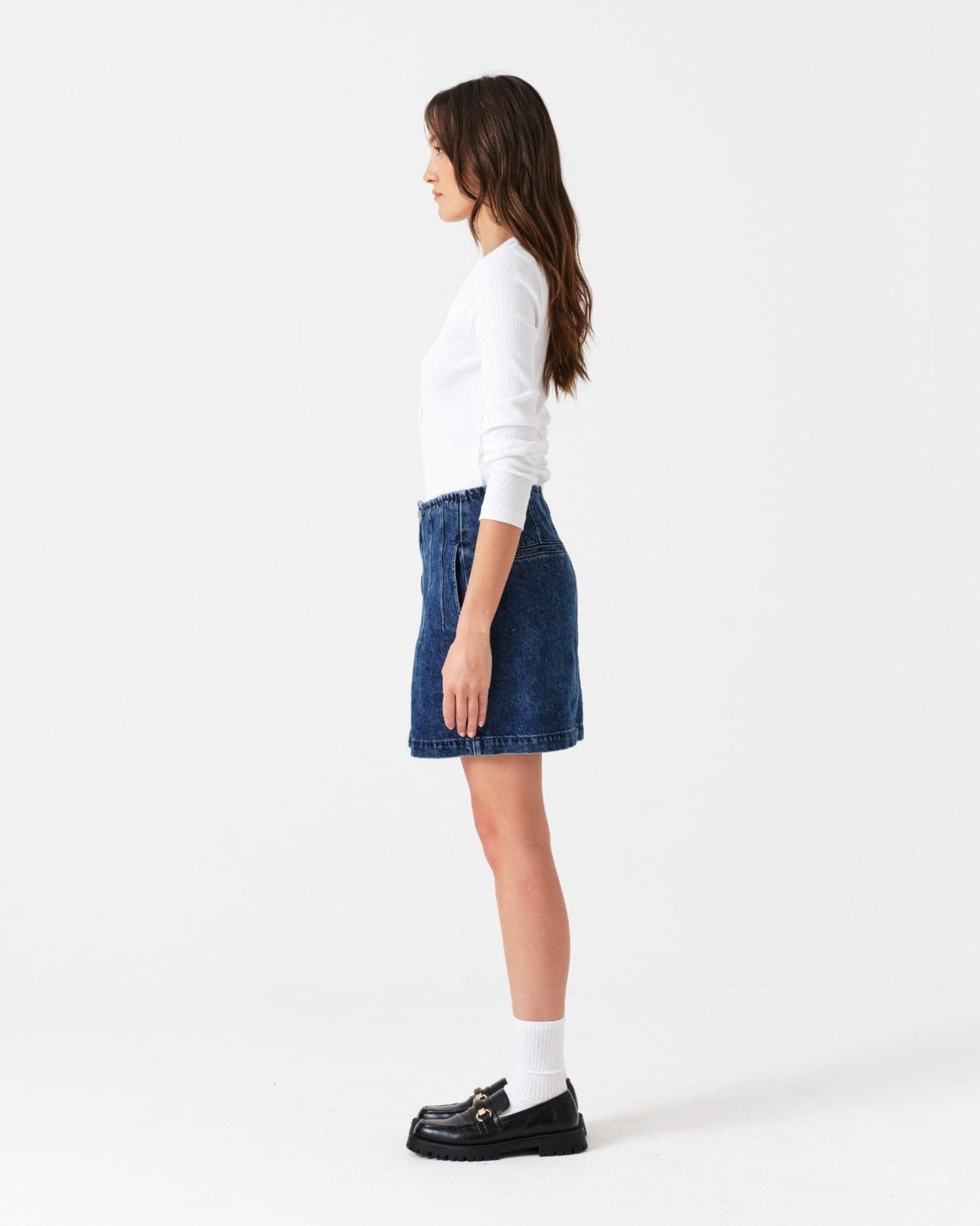 Person standing at the side and wearing a white sweater, blue mini denim skirt, and black shoes.