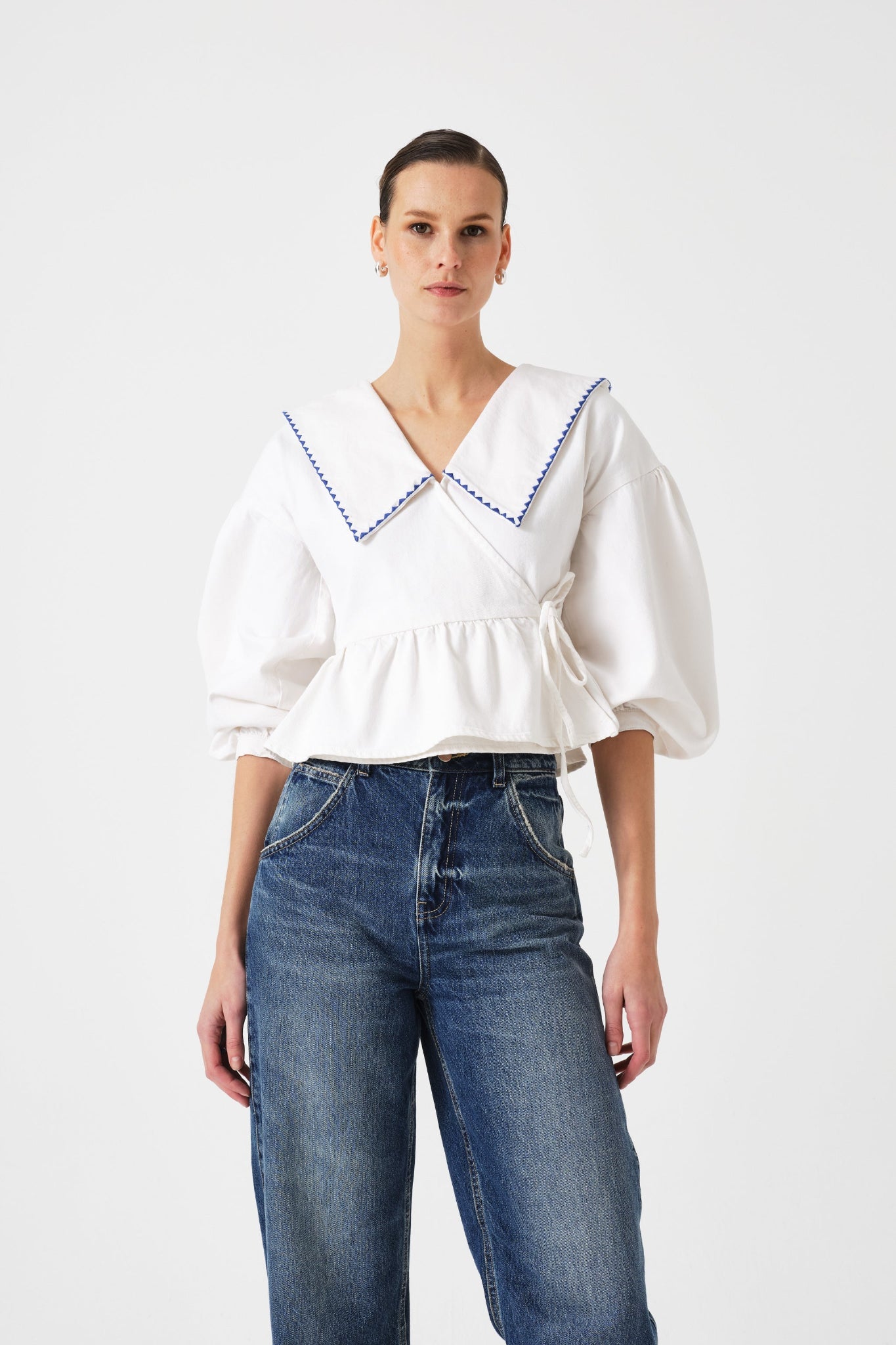 Woman wearing a white wrap blouse with blue trim on collar, sleeves pushed up and blue jeans