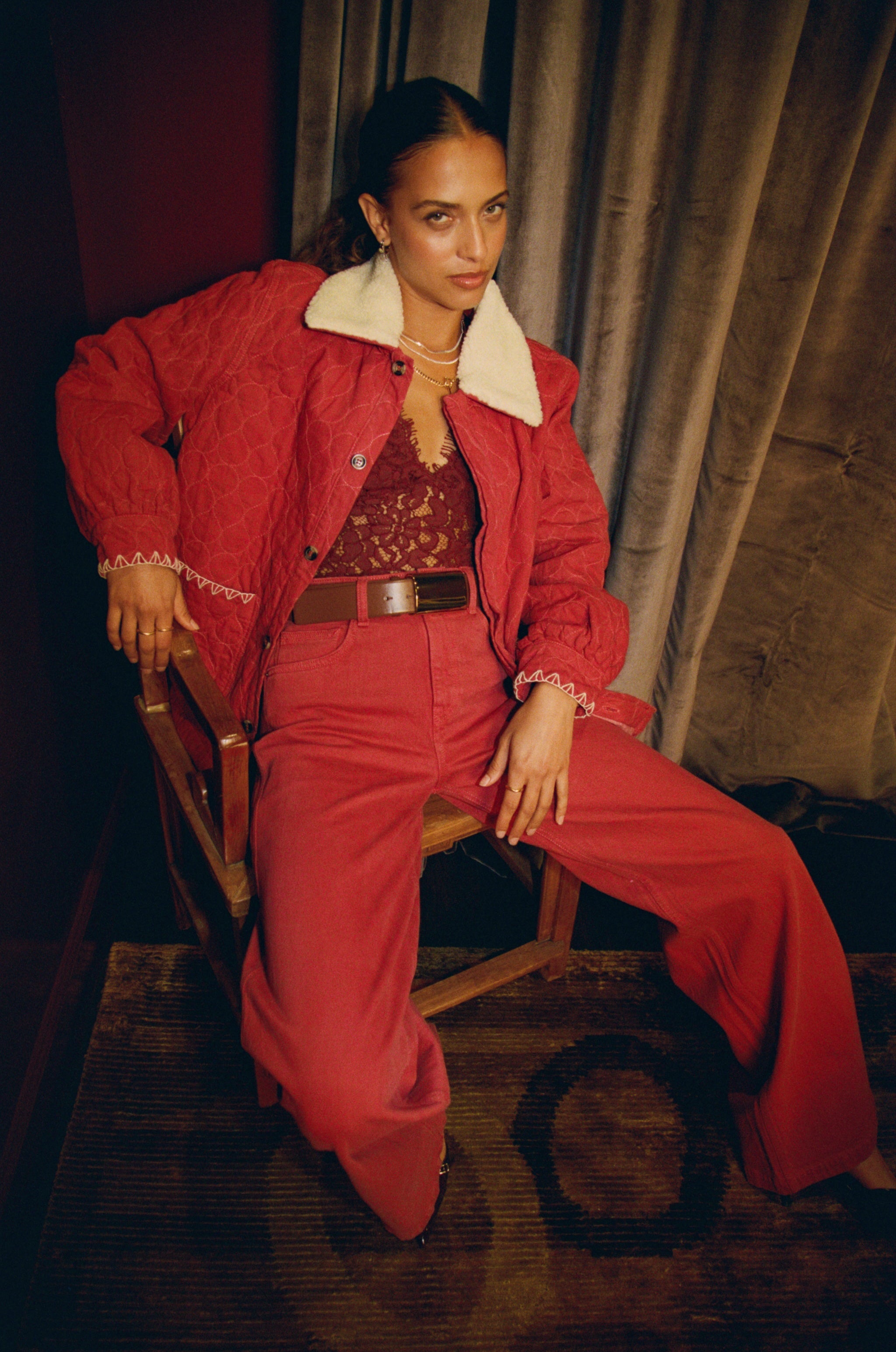 Woman in a faded rose jeans and jacket denim outfit sitting on a chair with a curtain background