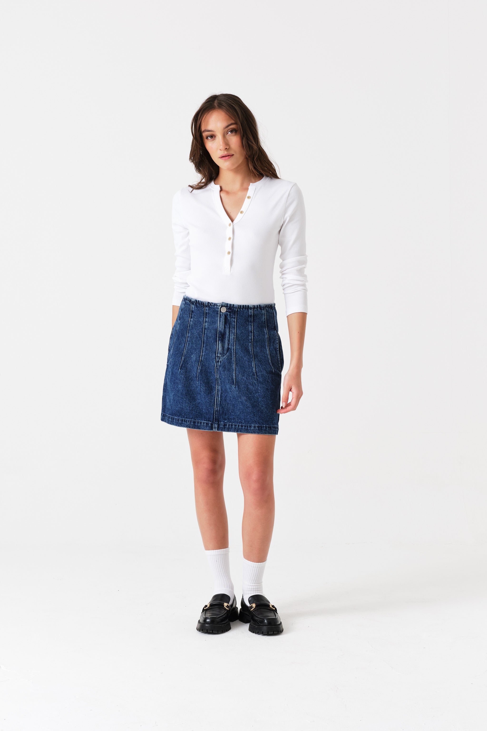 Woman wearing a white shirt, blue denim mini skirt with corset seam detailing in front.