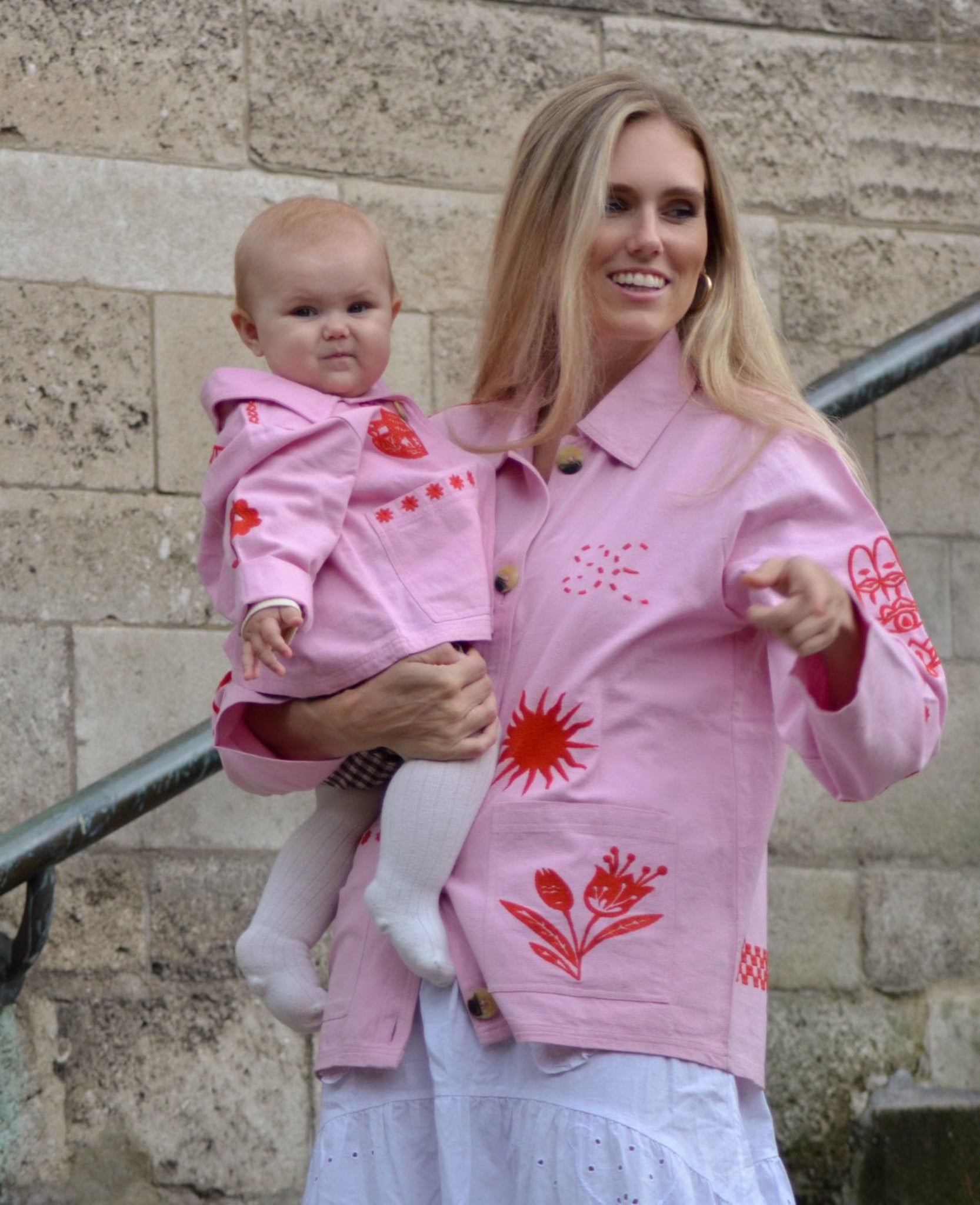 Mother holding a toddler in matching pink jackets with red embroidery.