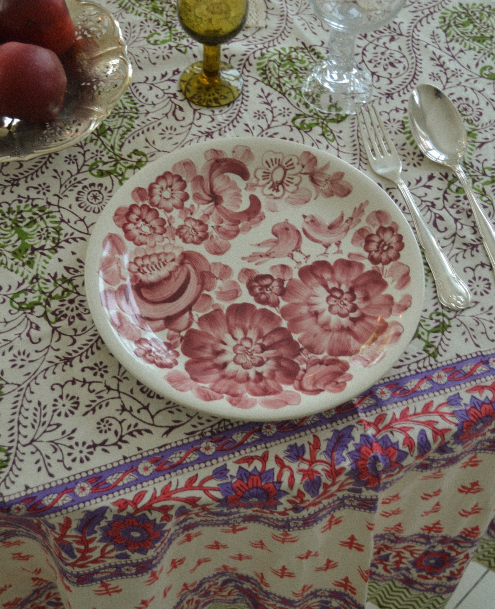 Close up of a green, and purple flower patterned cotton tablecloth on a table set with floral plate and silver cutlery.