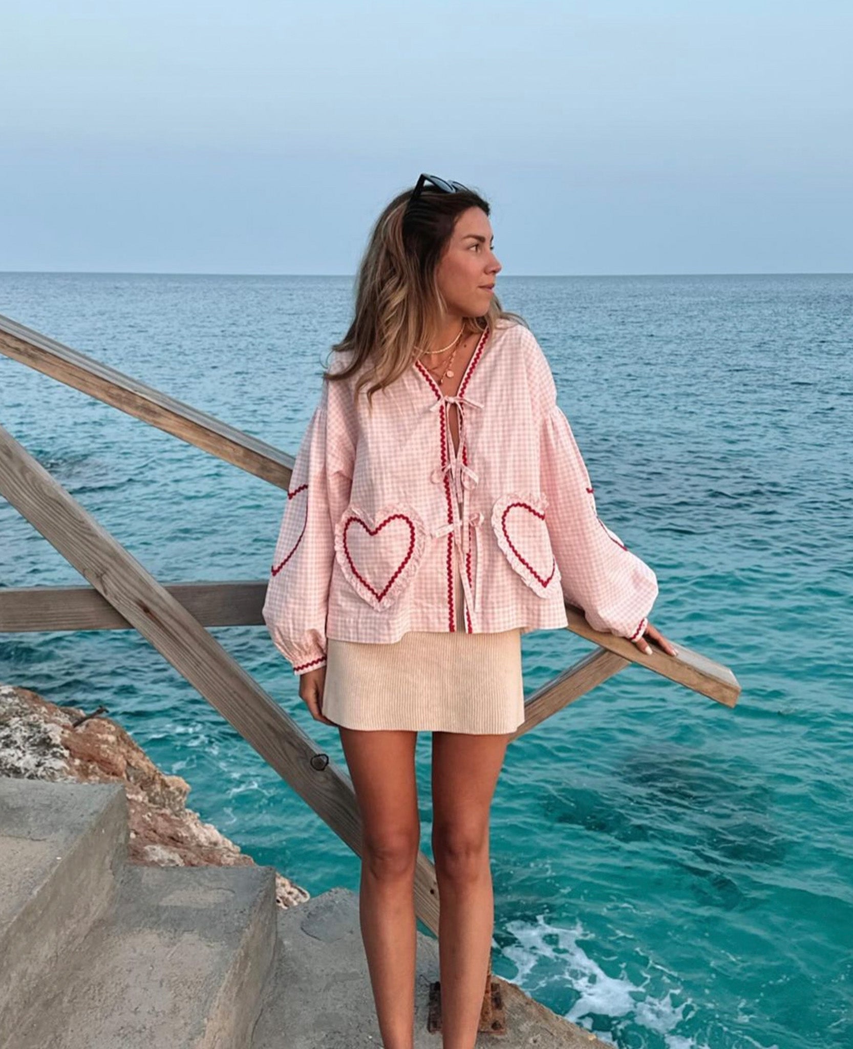 Woman standing on path near ocean wearing long sleeve pink checked blouse with front tie closures and heart shaped pockets and mini skirt.