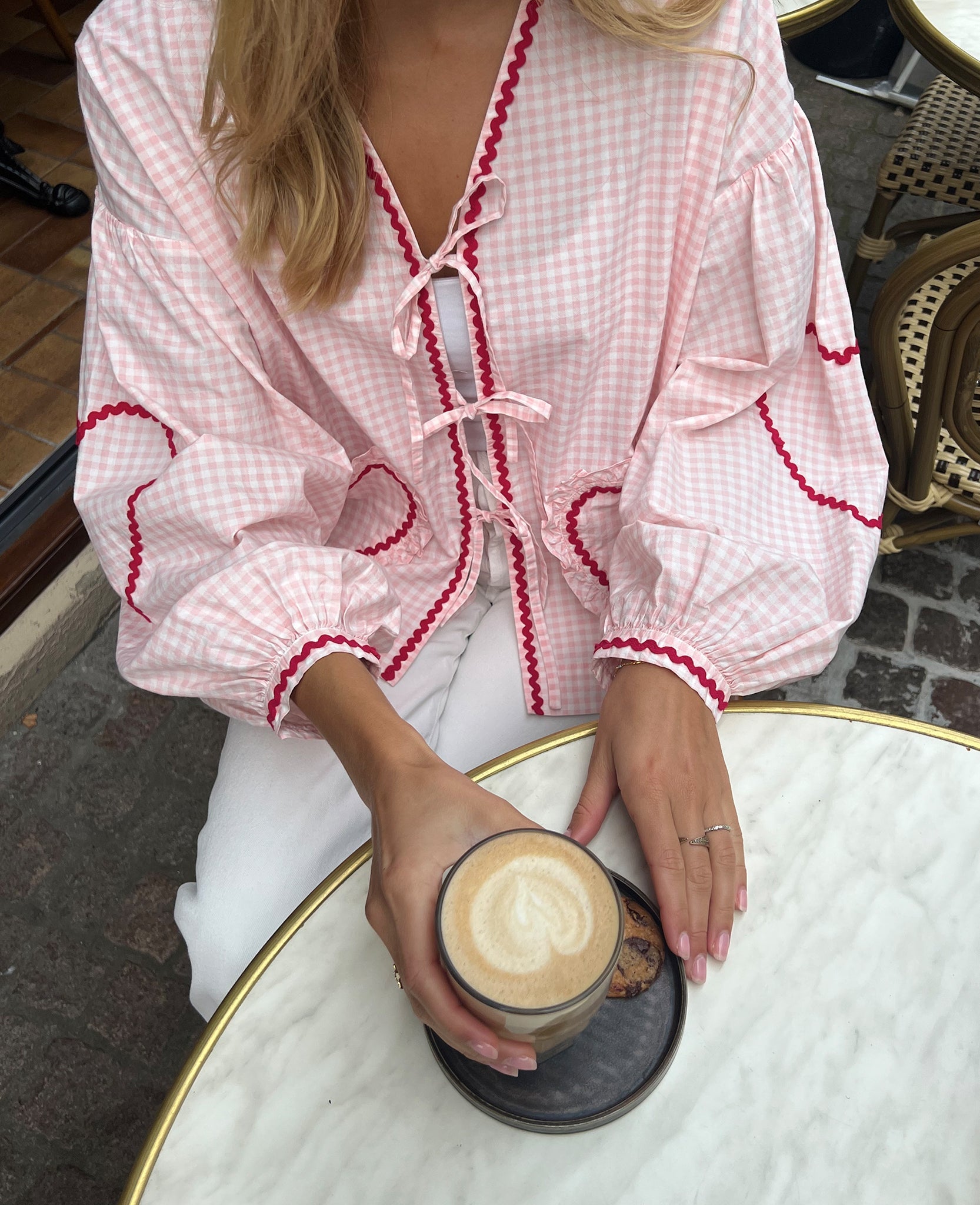Woman drinking coffee at cafe table wearing pink checked long sleeve blouse and tie closures over a white top and jeans.