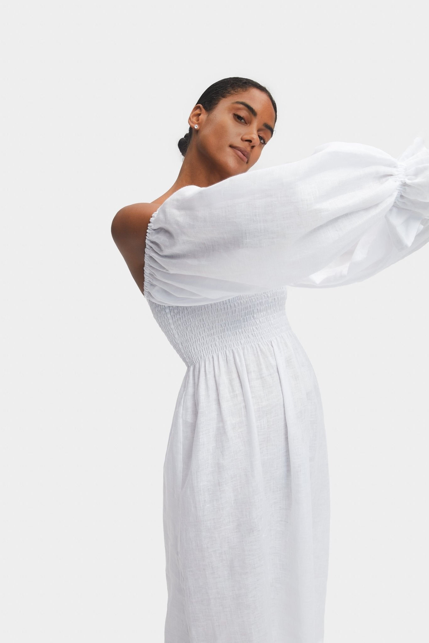 Woman standing on the side holding up her arms in the air wearing a white smocked bodice dress showing the puffed long sleeve.