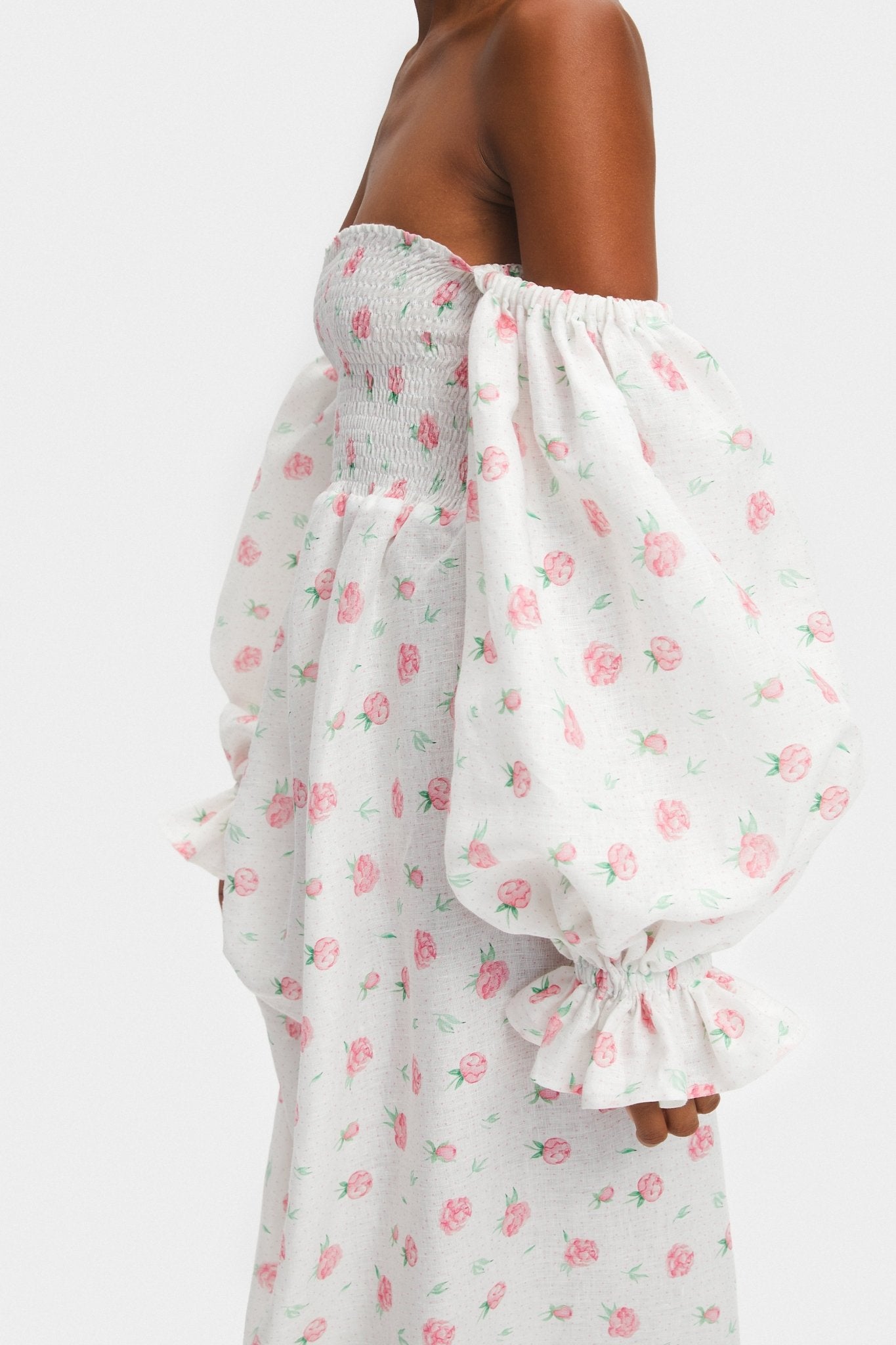 Side view of a woman wearing a white linen rose print dress with long sleeves off the shoulder and shirred bodice.