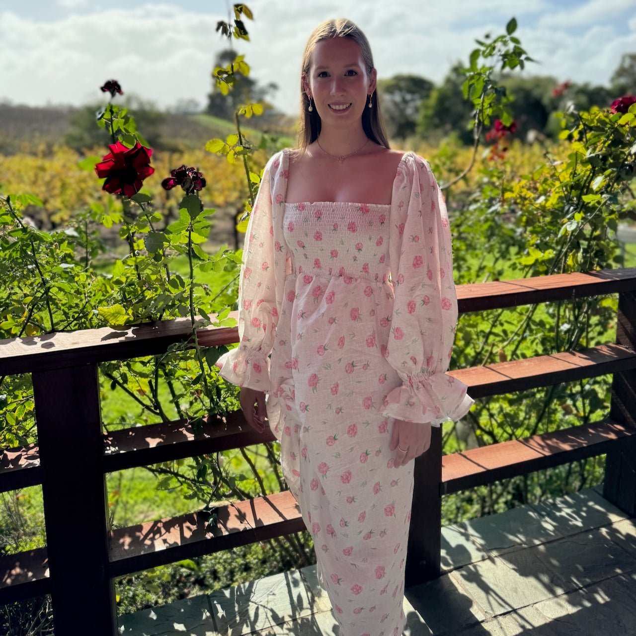 Woman in the garden wearing a white linen dress with rose print pattern, long sleeves on the shoulder with elasticated cuffs.