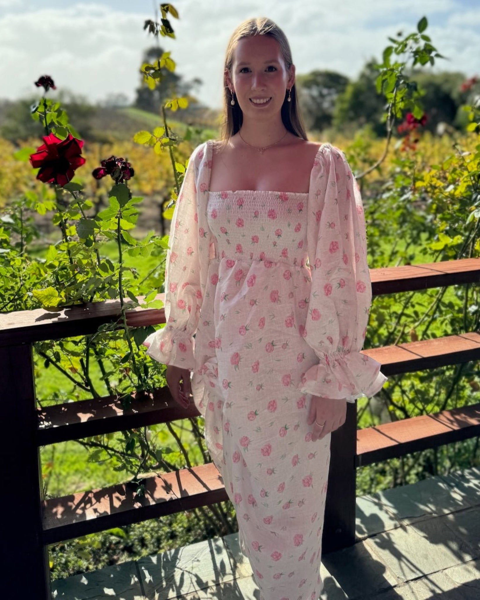 Woman in the garden wearing a white linen dress with rose print pattern, long sleeves on the shoulder with elasticated cuffs.