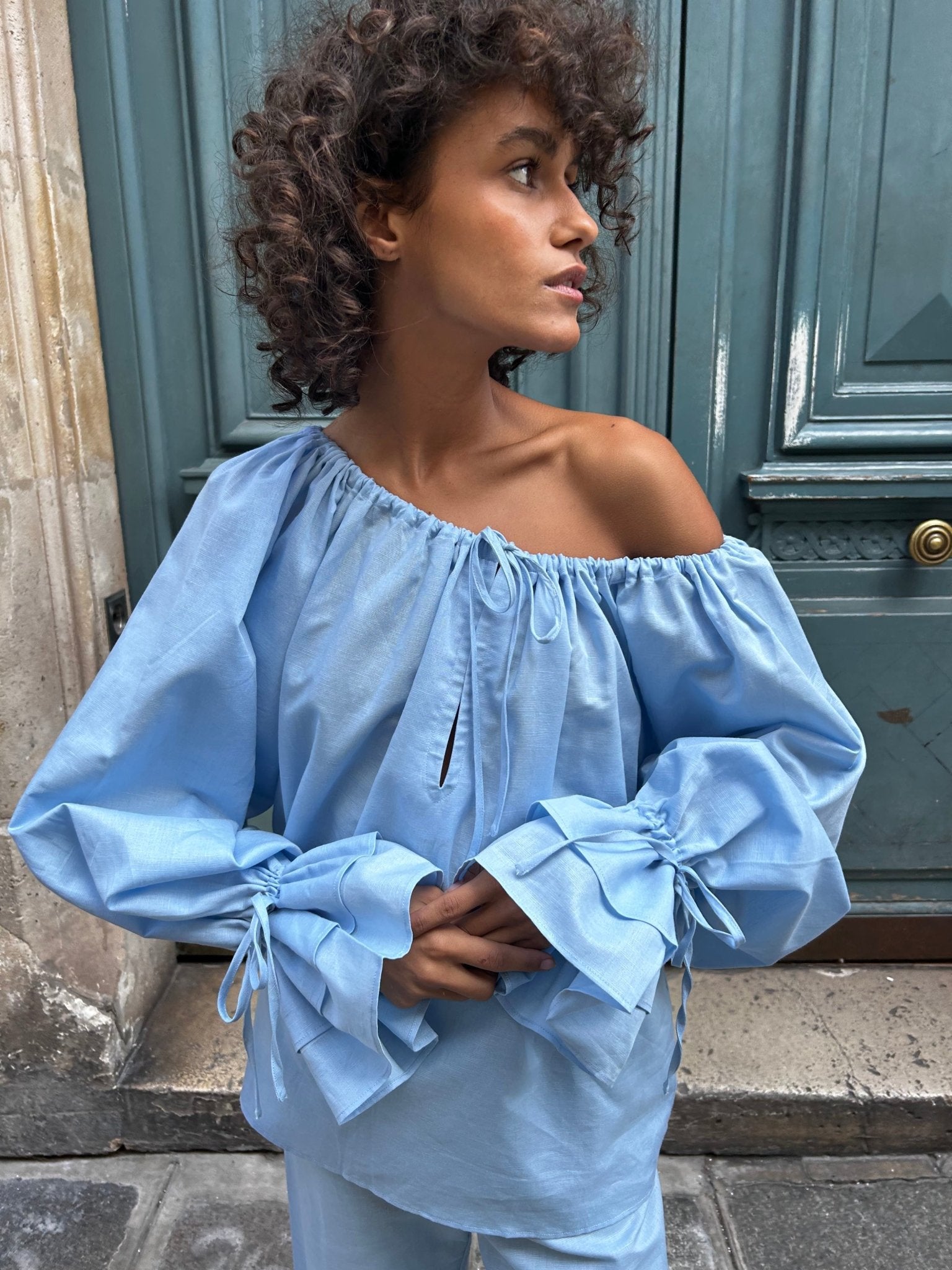 woman-wearing-a-linen-blue-blouse-with-long-sleeves-and-adjustable-round-neck-with-ties-worn-off-the-shoulder-and-pants-in-blue