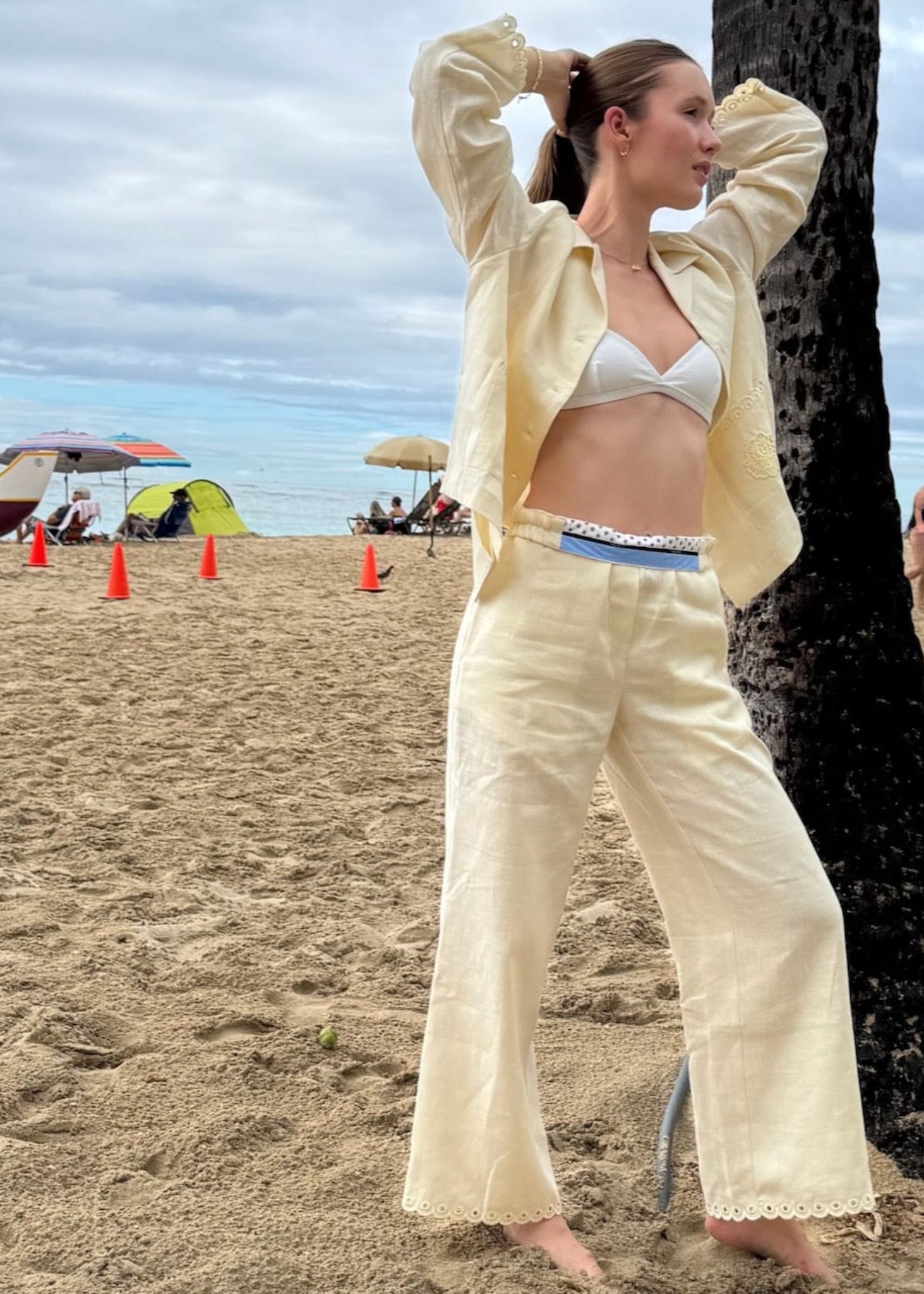 Woman in a cream linen shirt and pants standing on a beach with palm trees and umbrellas in the background.
