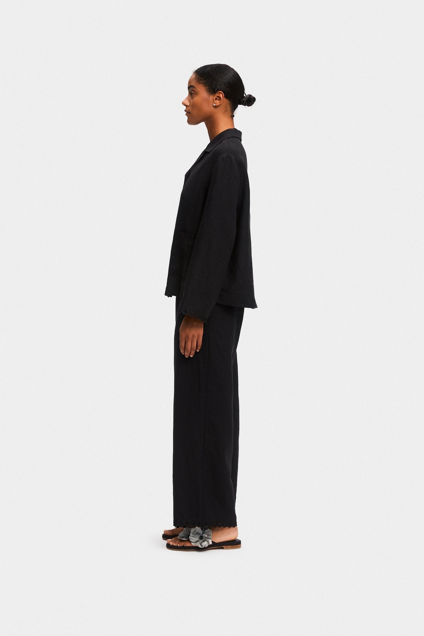 Woman standing on the side wearing black shirt and pants with scalloped embroidered hem and black shoes.