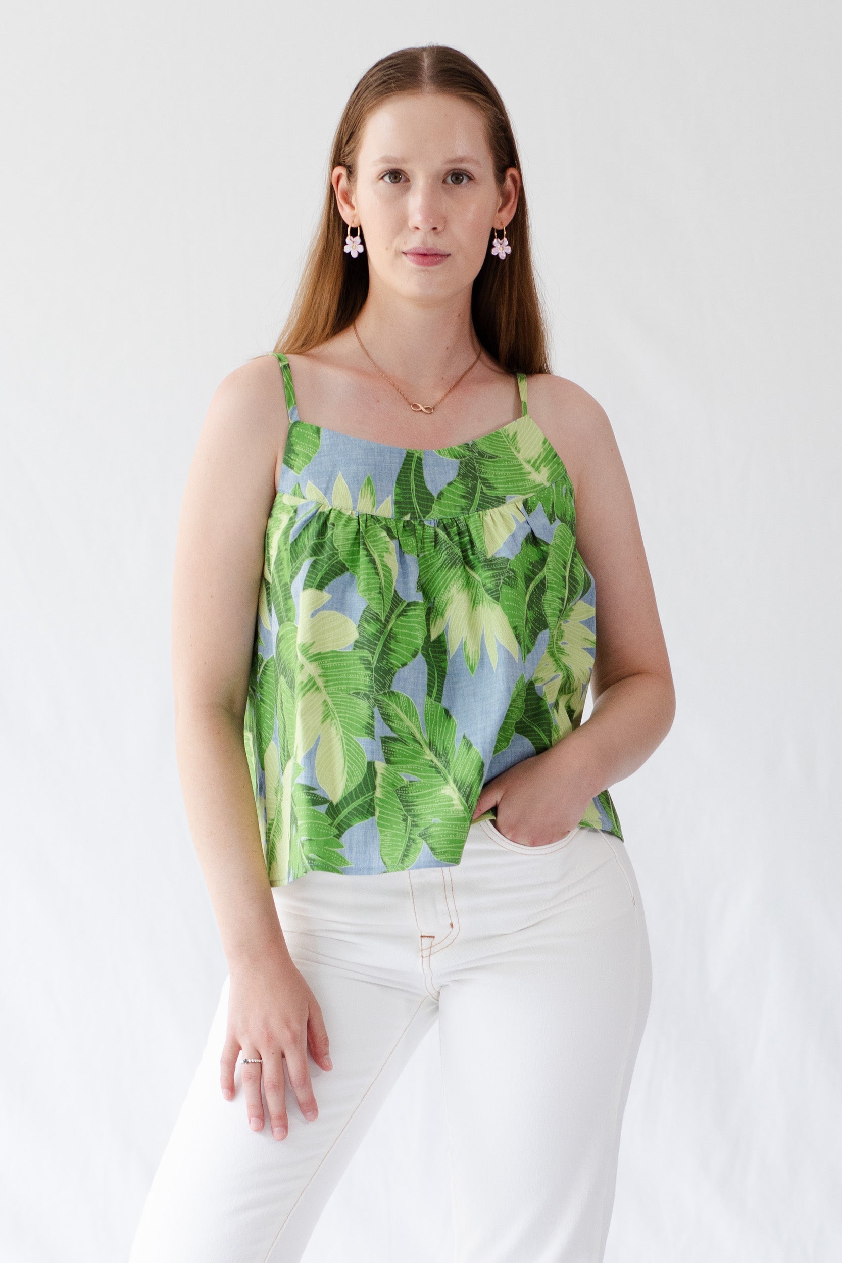 girl-wearing-Ariel-cami-top-with-tie-straps-QLD-fabric-large-green-leaves-on-light-denim coloured-background-and-white-jeans-facing-front