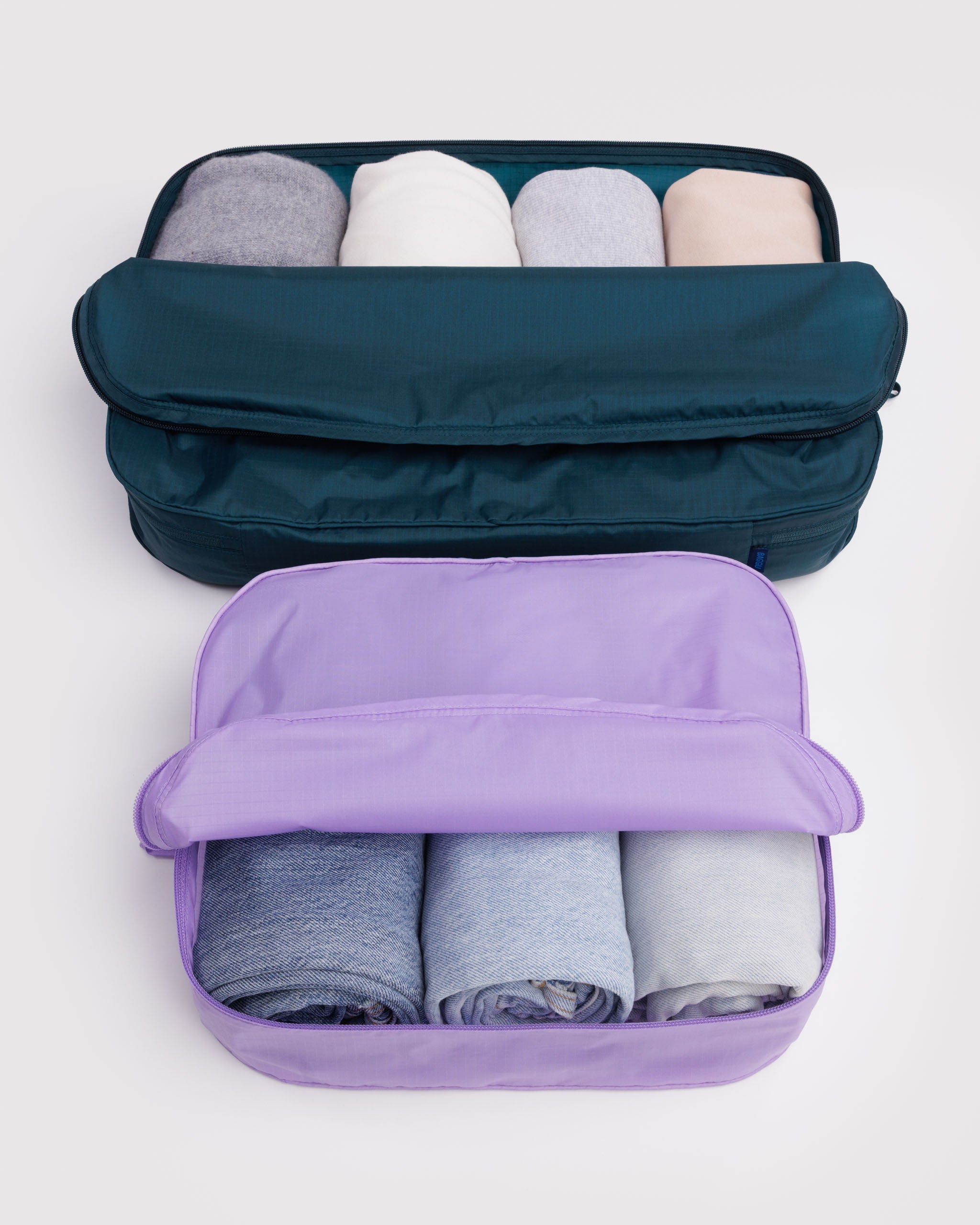 Two different sized packing cubes, one medium teal and one small purple, filled with rolled-up clothes and zip top folded back in half.