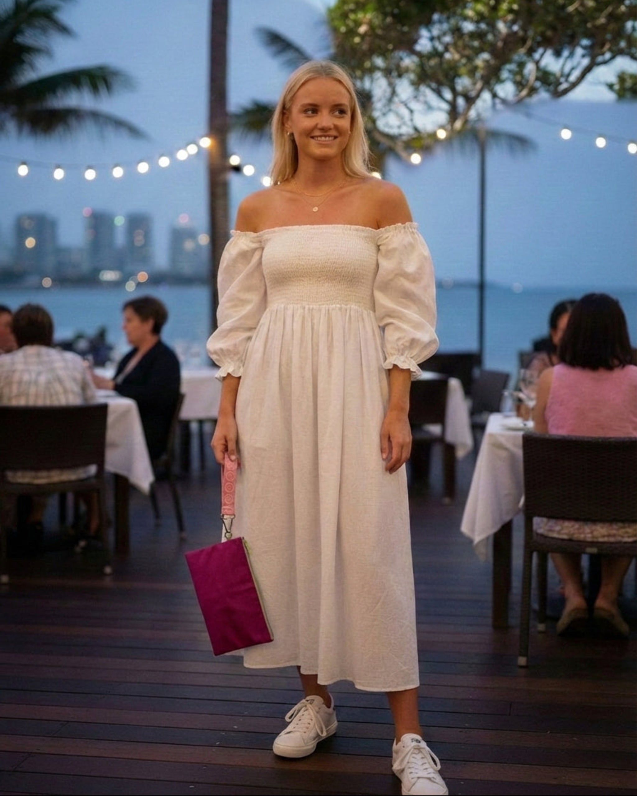Woman in a white off-shoulder dress standing in an outdoor dining area holding a pink logo keychain attached to a medium go pouch as an evening clutch.