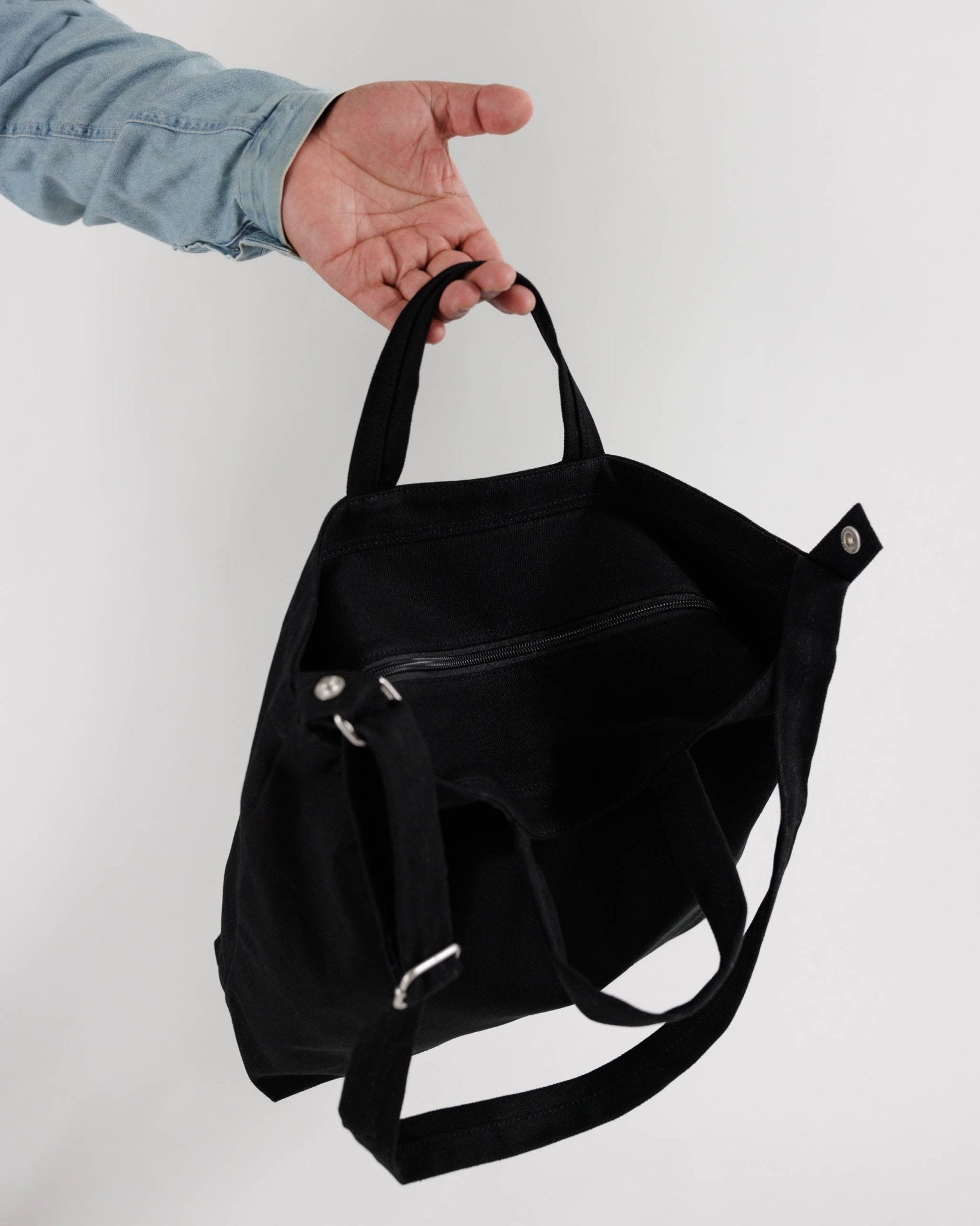 Black horizontal tote with a zip closure and an adjustable shoulder strap held by a person by the short handle.