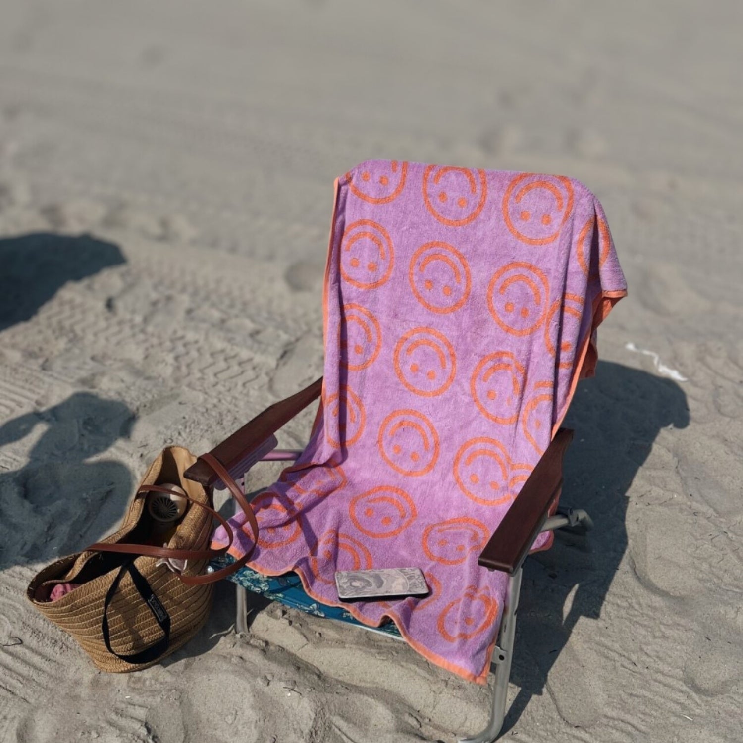 Beach chair with a purple towel featuring orange smiley face patterns on a sandy beach.