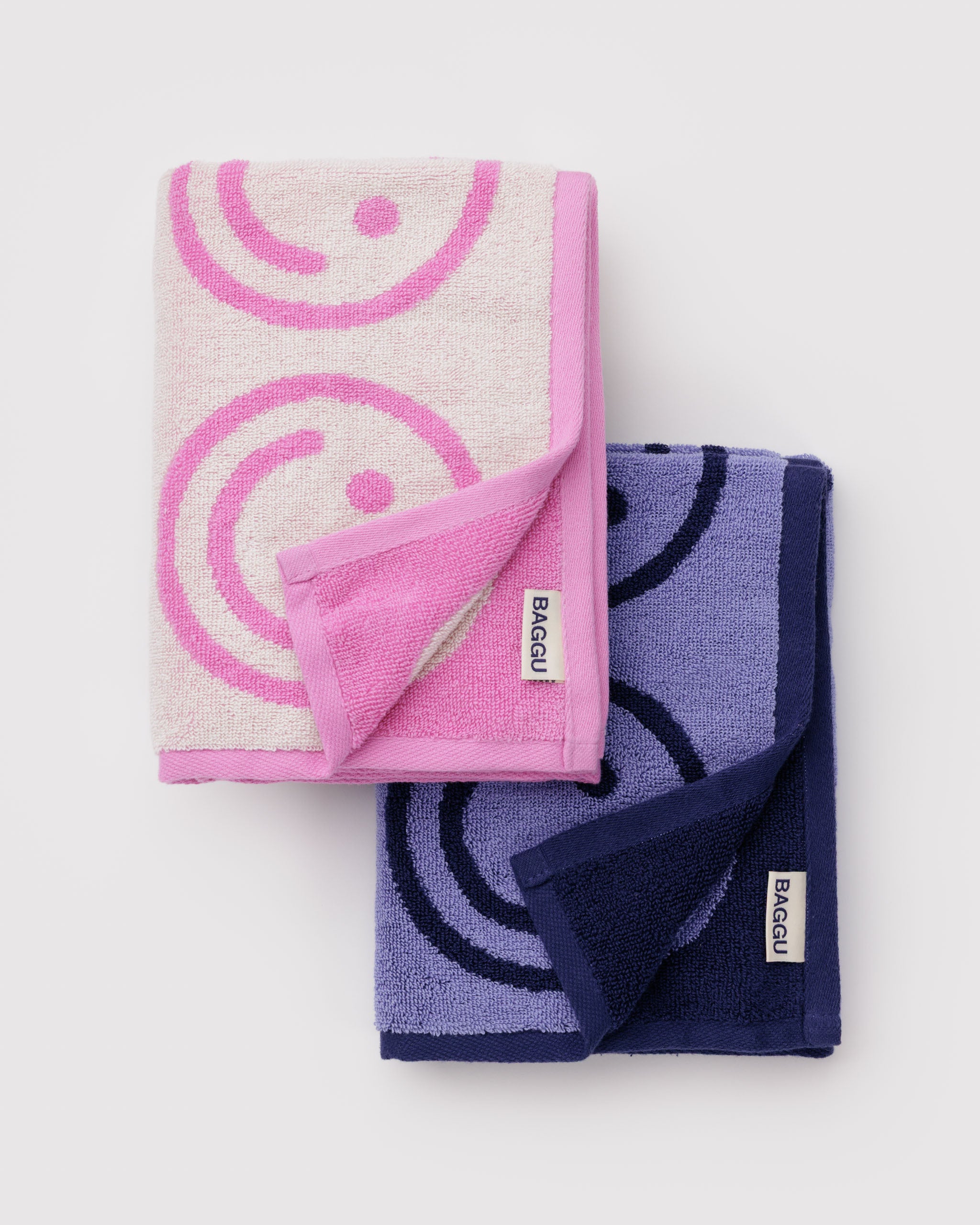 Two folded towels, one pink with a circle smiley face pattern and the other blue with baggu logo tag.