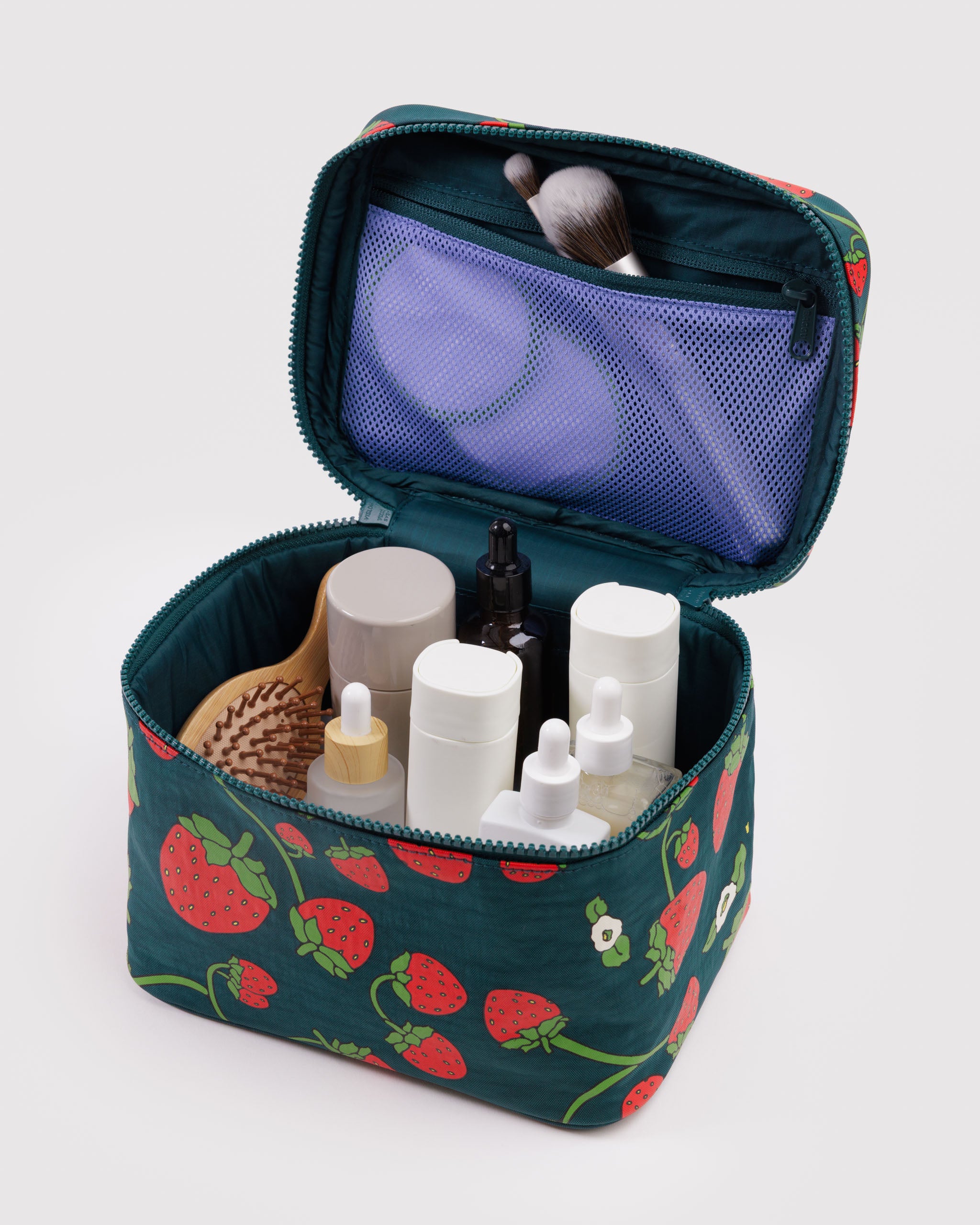 Open green cosmetic bag with strawberry and flowers pattern containing various skincare products and makeup brushes in zip mesh pouch on lid.