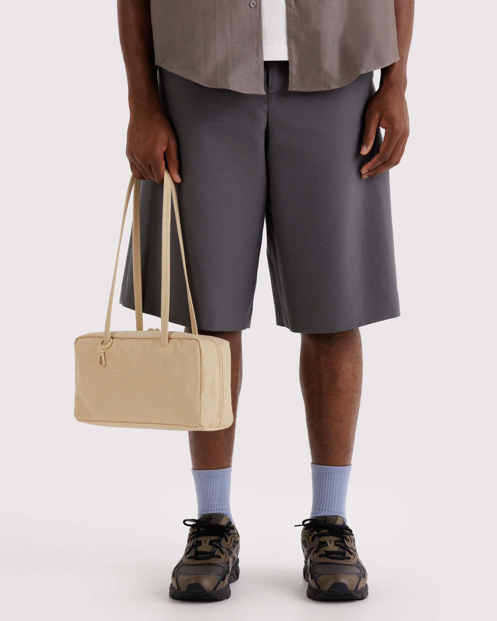 Male person holding long double handles of a light yellow handbag wearing grey shorts and brown shoes.