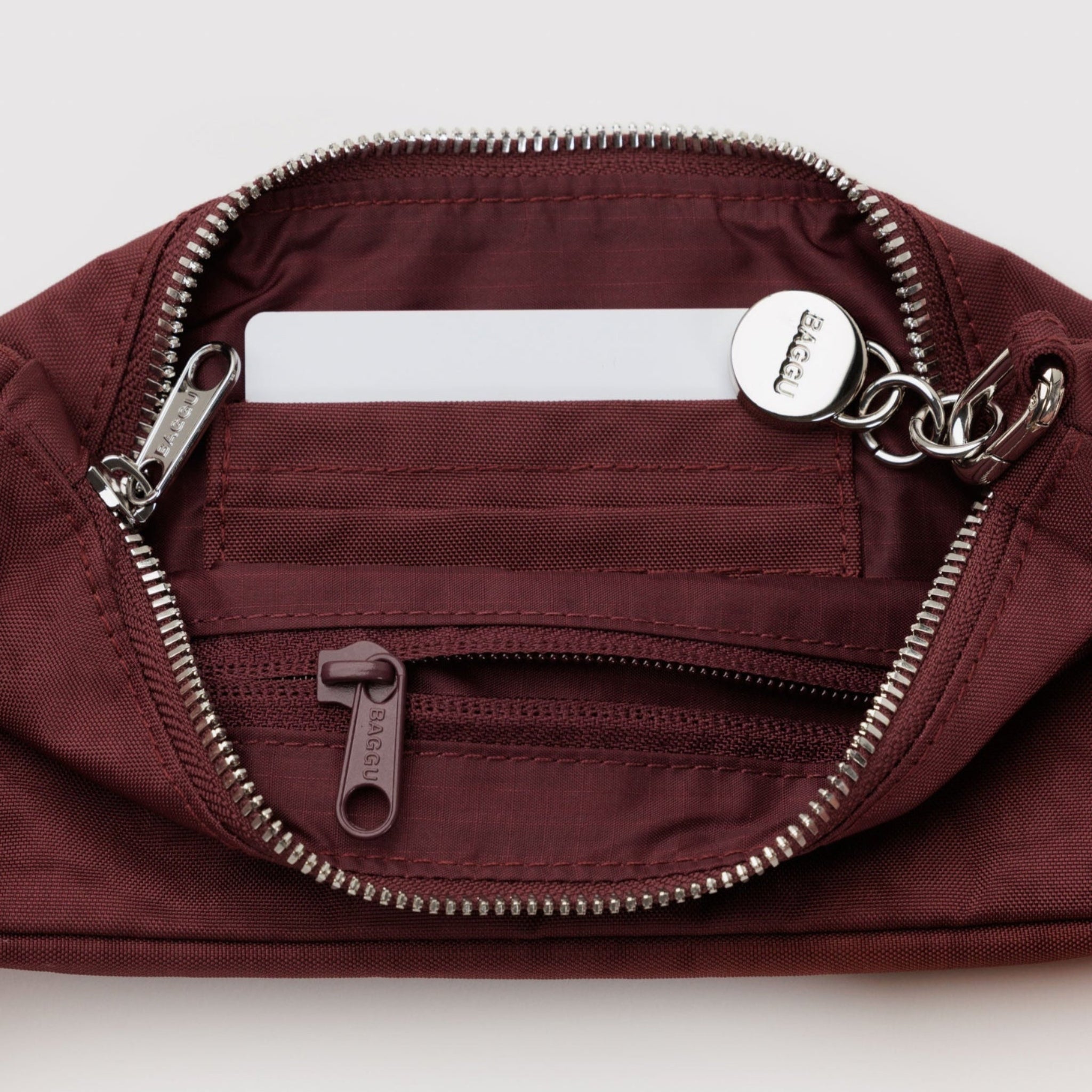 Mahogany bag with top silver zipper showing internal tonal zip pocket and card slots with adjustable metal clasp for shoulder strap and BAGGU branded charm.