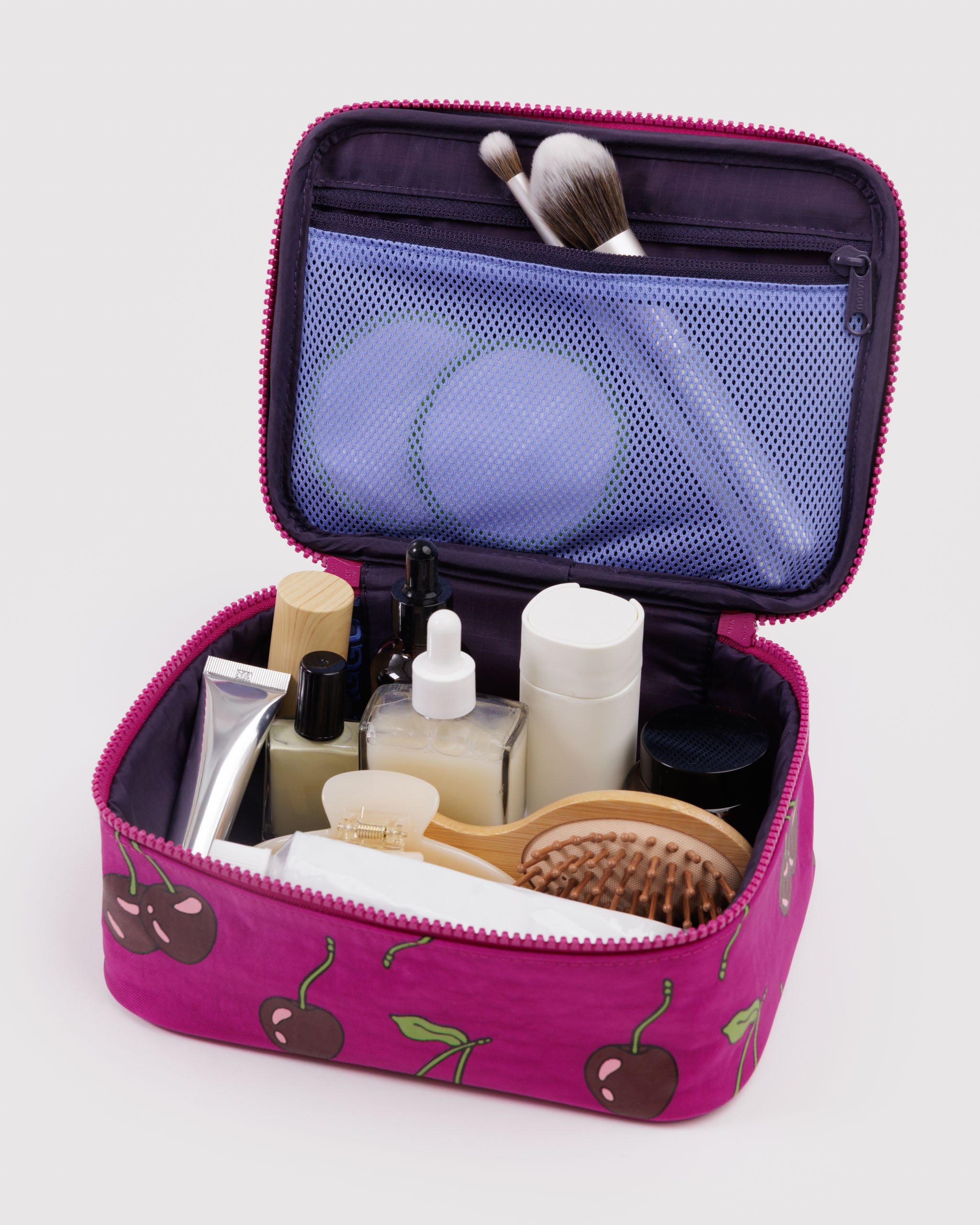 Pink cosmetic bag with cherrries pattern holding toiletries inside and makeup brushes stored in mesh zip pocket on lid.