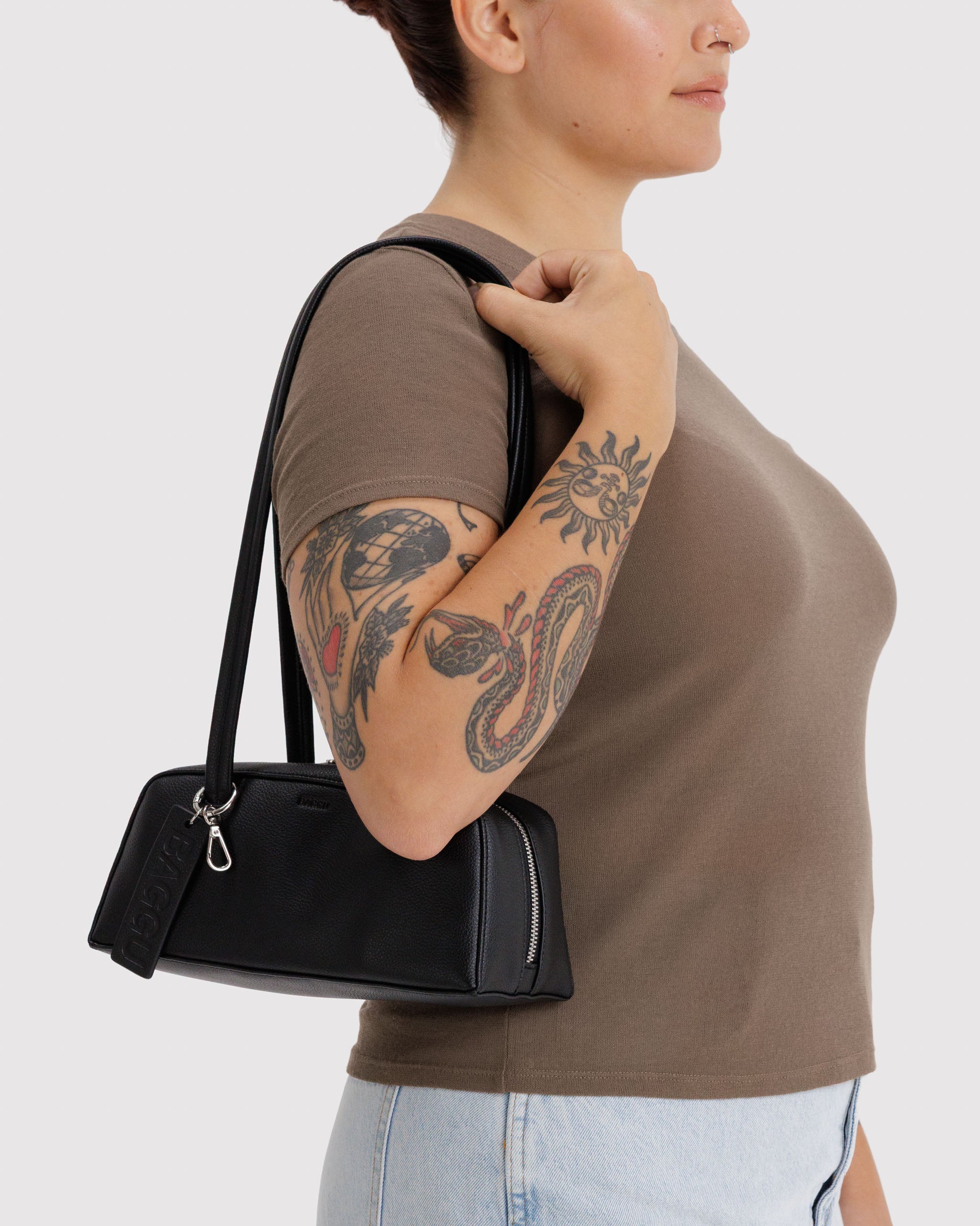 Person wearing a brown shirt holding a black leather handbag with metal top zip, a metal clip and BAGGU leather tag on one of the double shoulder straps.