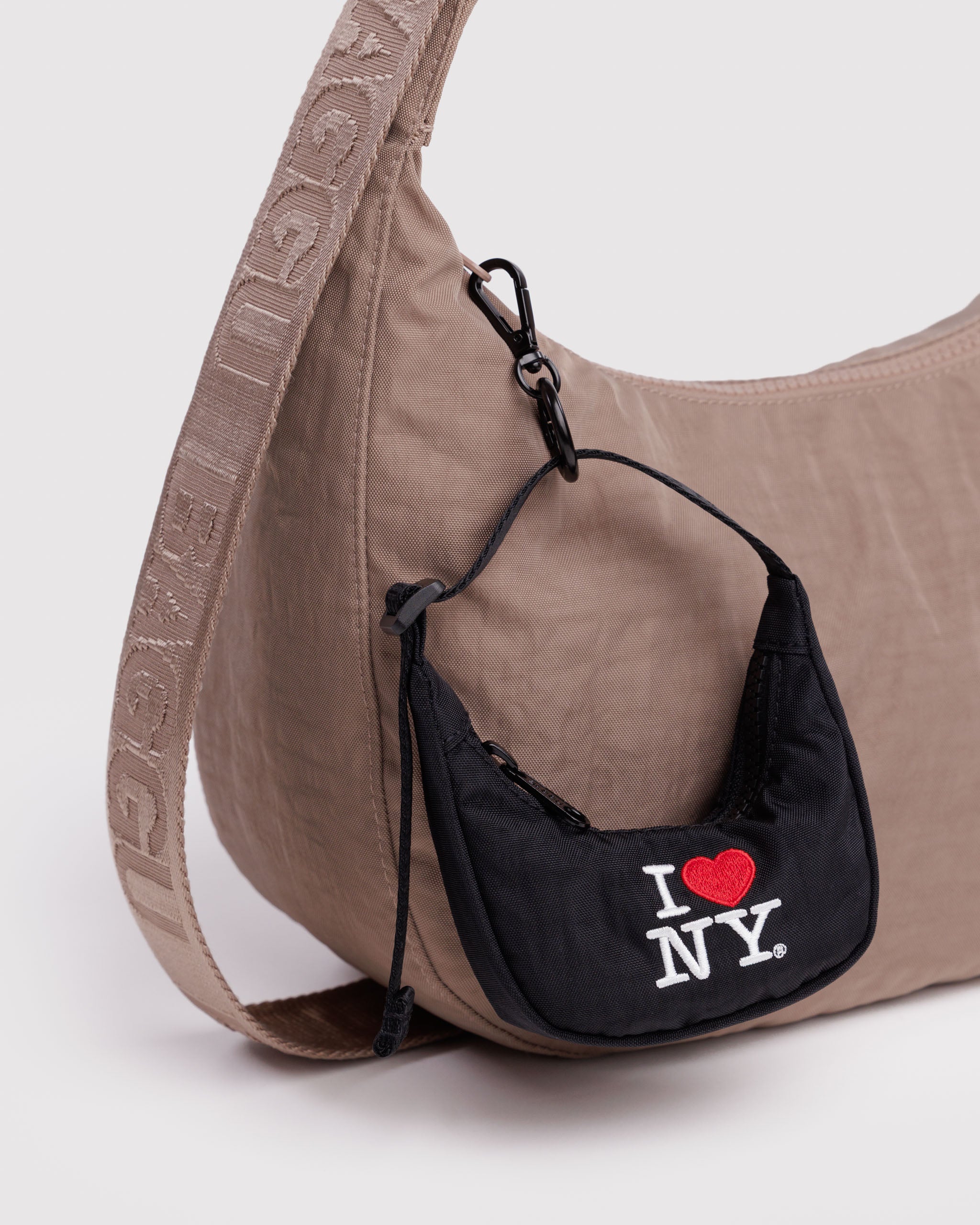 Brown handbag with a smaller black crescent shaped bag charm attached featuring 'I ❤️ NY' embroidery and a black clip.
