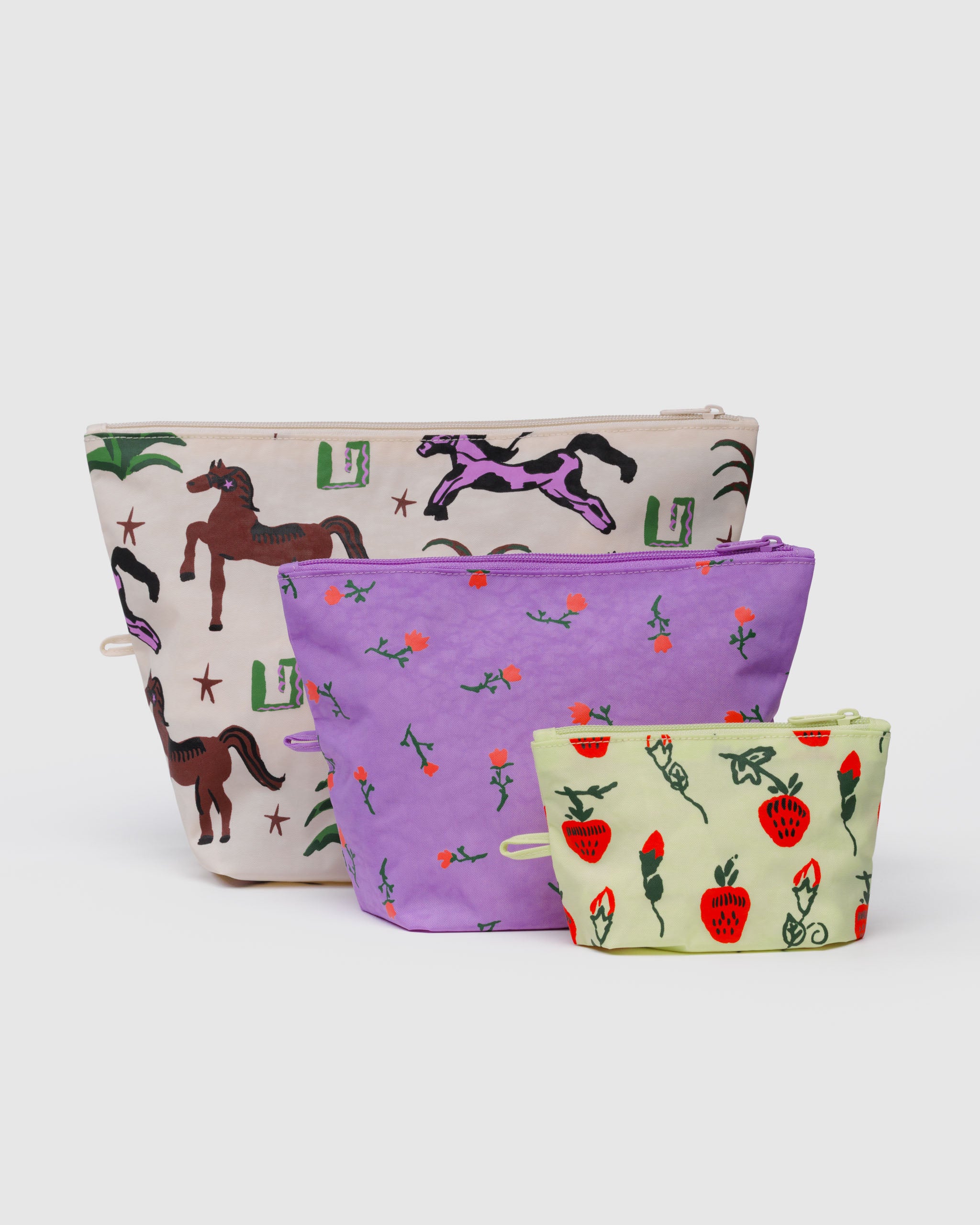 Three fabric pouches with zip closure in different patterns of horse, wildflowers and strawberry with matching hanging loops at the side standing upright with expanded bases.