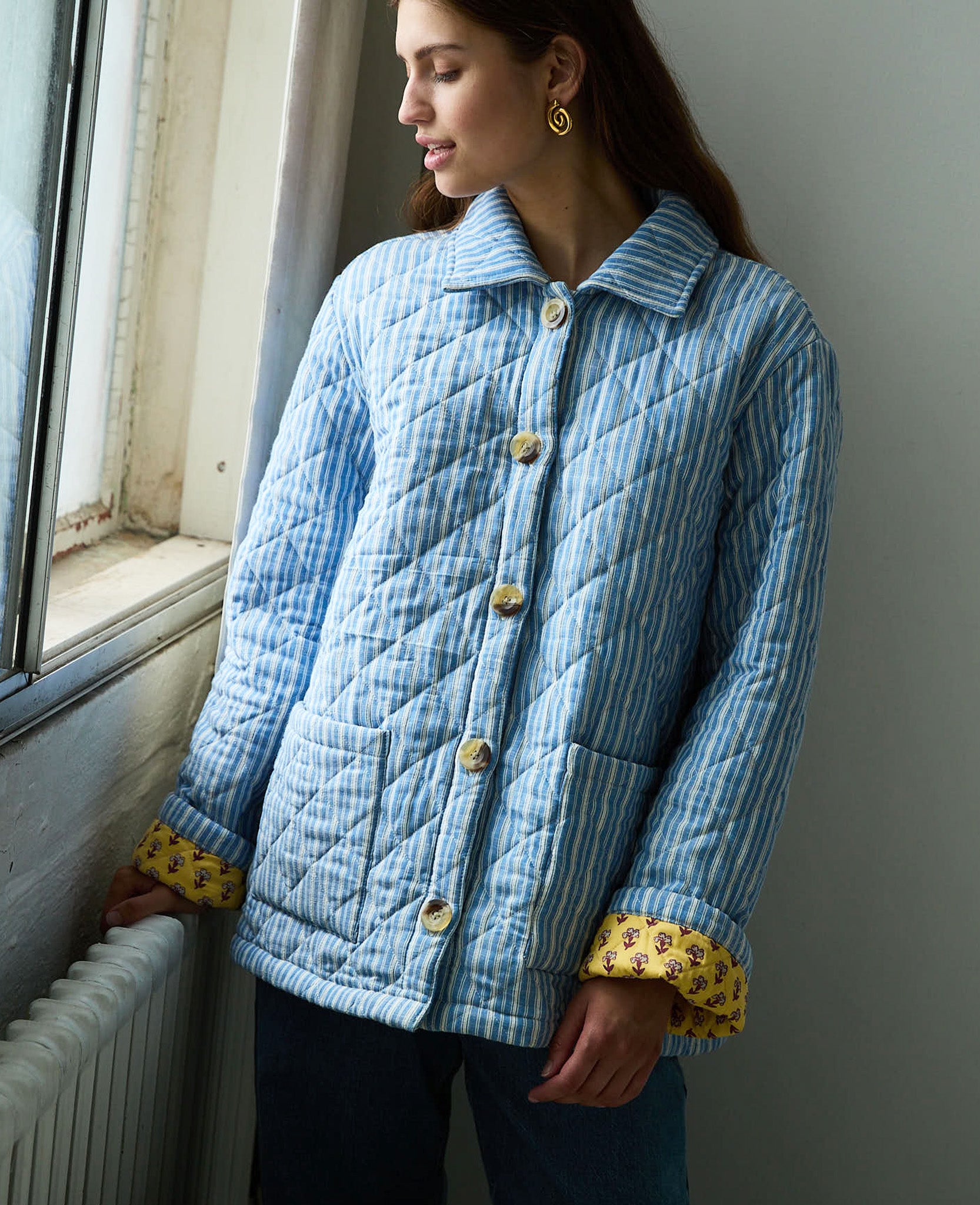 woman-standing-at -window-and-wearing-blue-and-white-striped-quilted-jacket-with-collar-and-yellow-floral-lining-buttons-done-up-two-patch-pockets