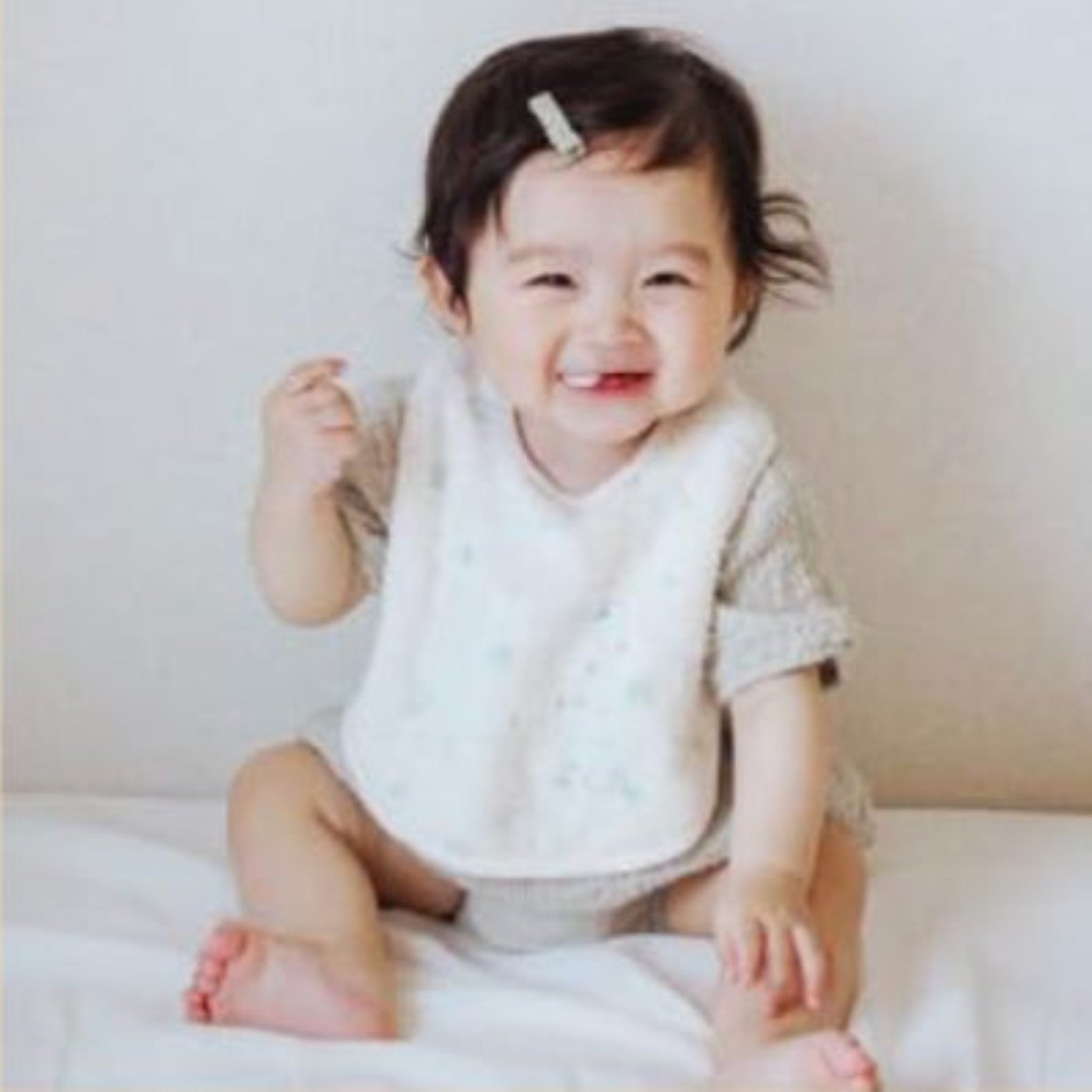 Baby sitting on white mat and wearing a cream apron bib with mint blue dots and small dog embroidery in bottom corner.