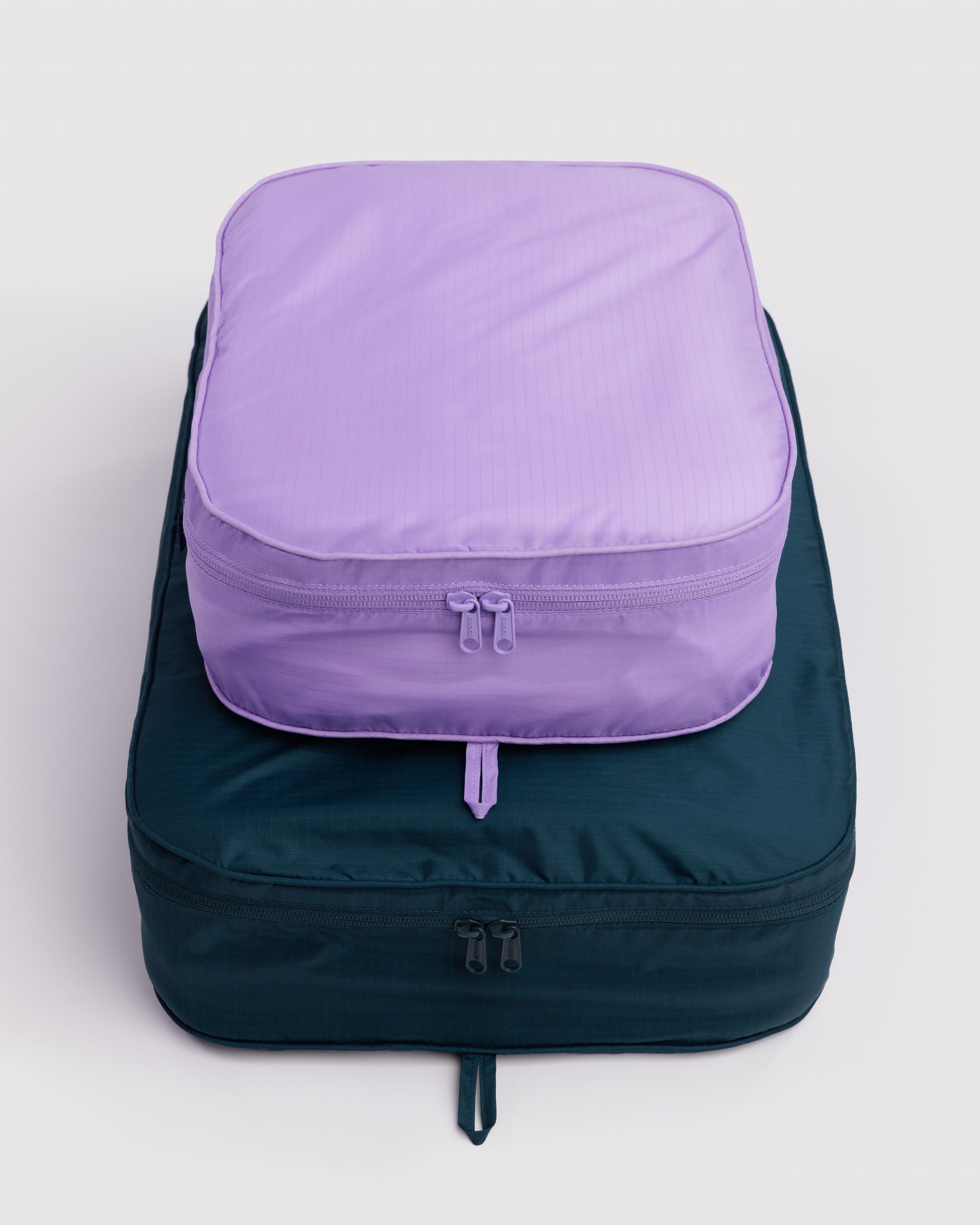 Two filled and stacked travel packing cubes, one purple and one teal, with double zippers and hanging loop.