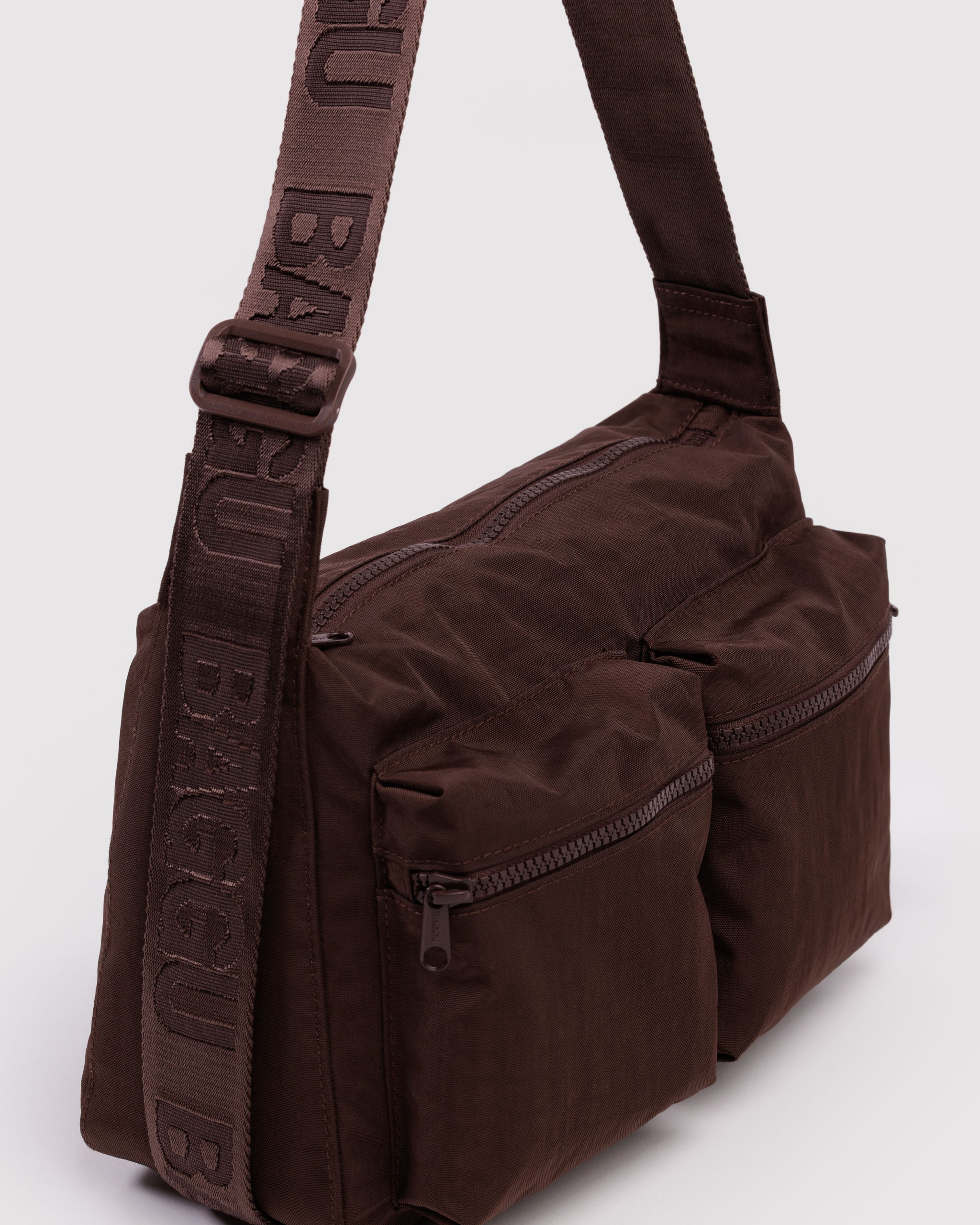 Brown shoulder bag with visible branding on wide webbed adjustable strap, top zip closure and two front zip cargo pockets.