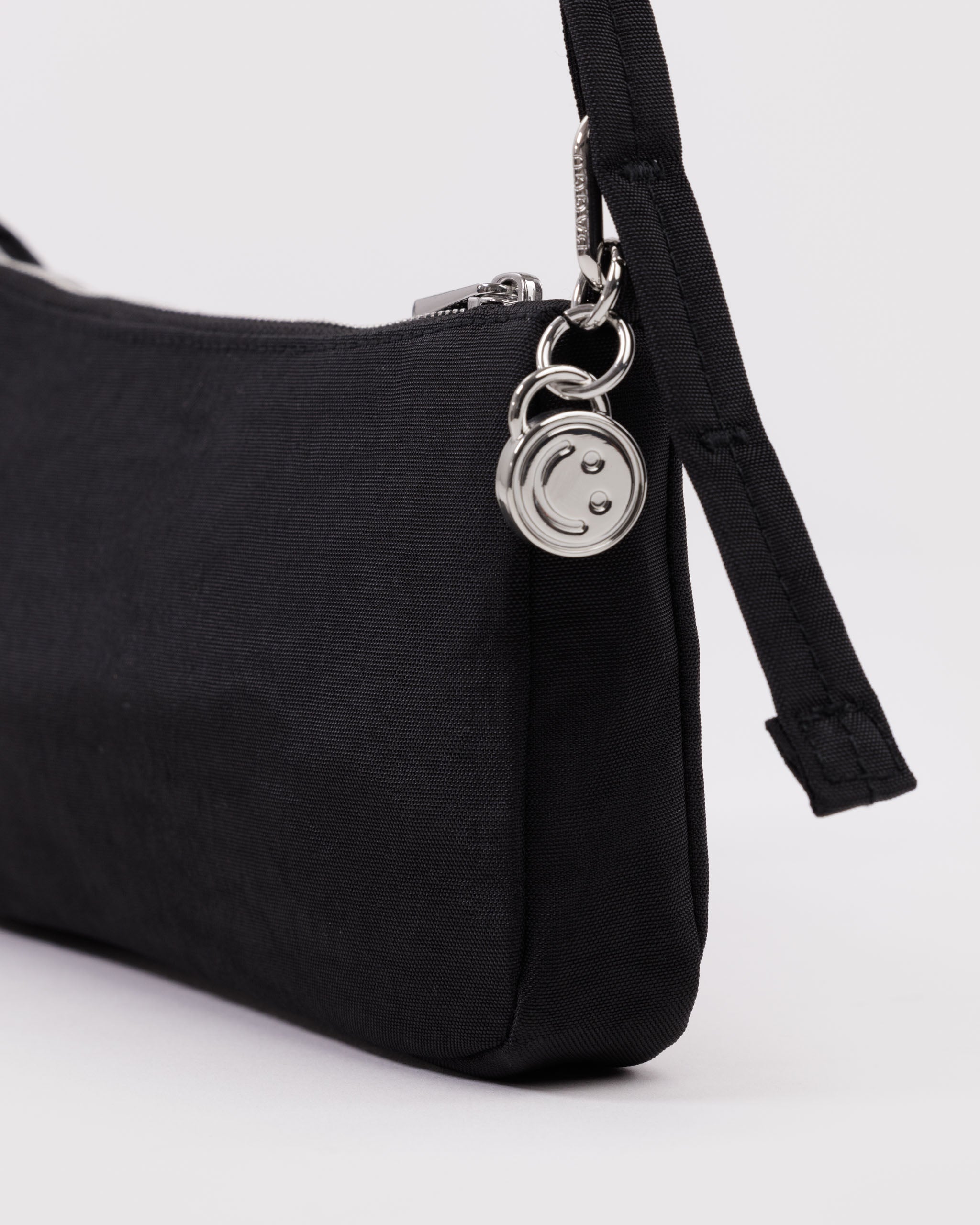 Black handbag with a metal silver smiley face charm attached to metal clasp to adjust the shoulder strap and  a metal zipper.