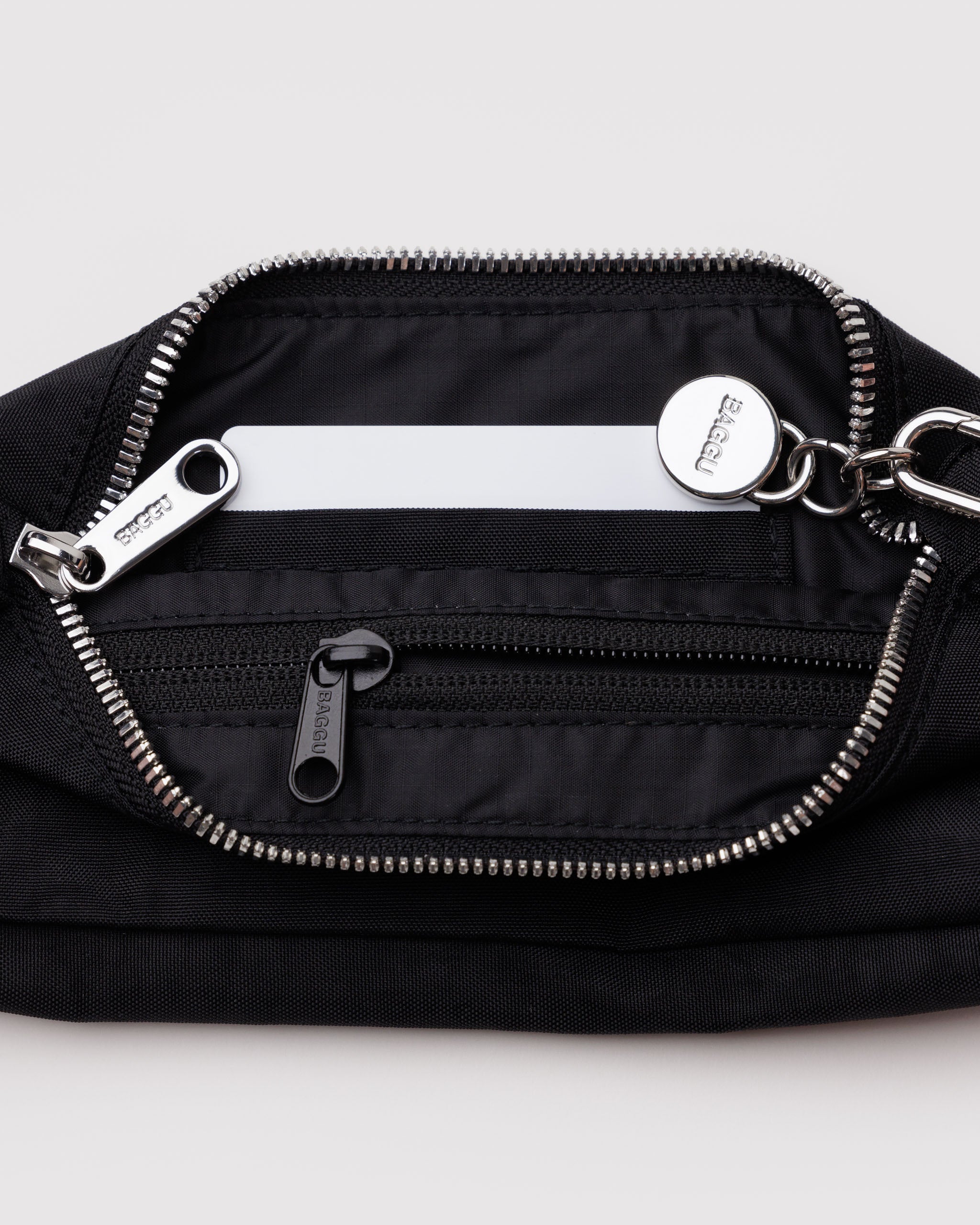 Top metal zip open on a black handbag showing internal tonal zip pocket, card slots and BAGGU branded metal charm on metal clasp.
