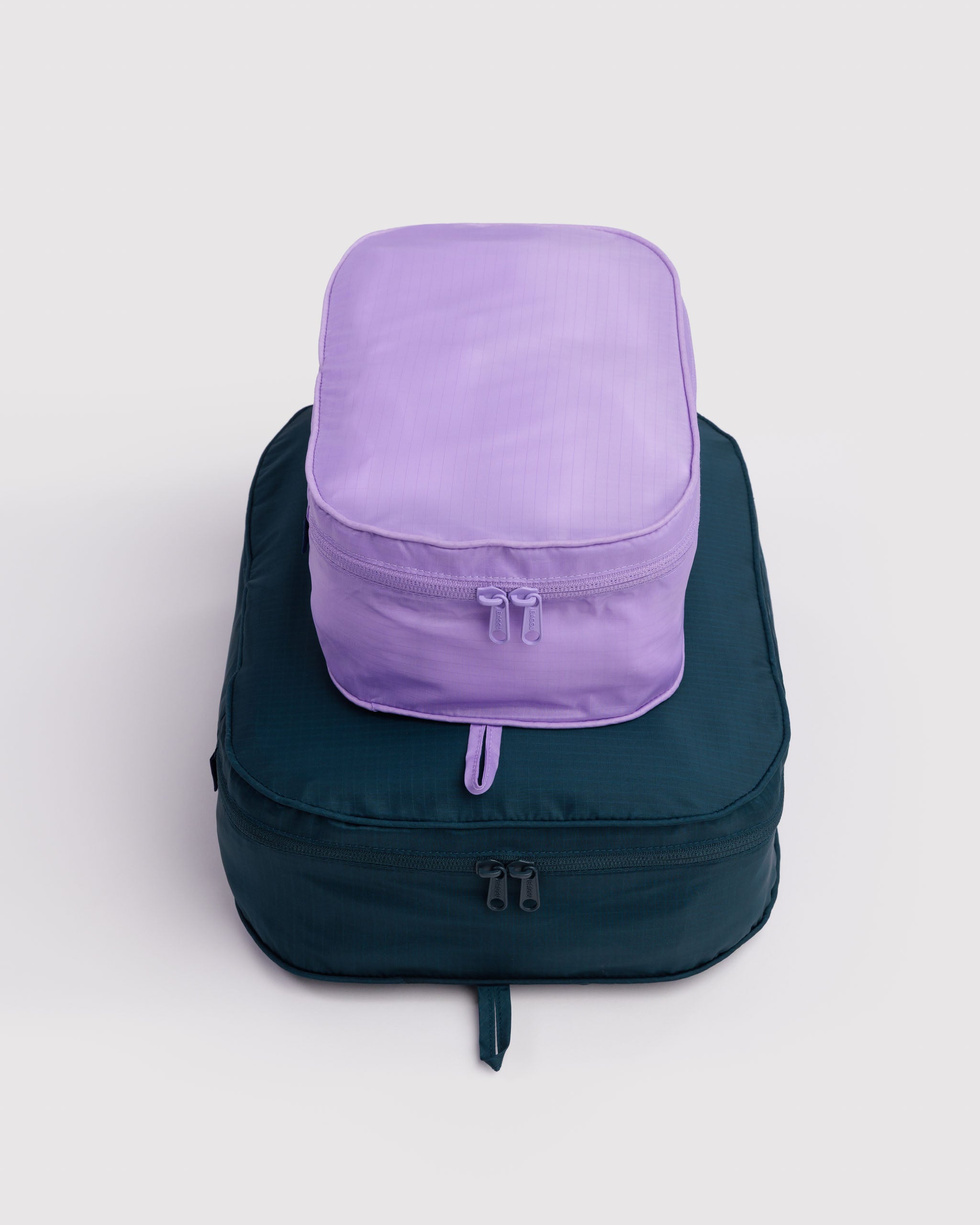 A filled purple packing cube-shaped bag on top of a teal packing cube with closed double zips and hanging loop.