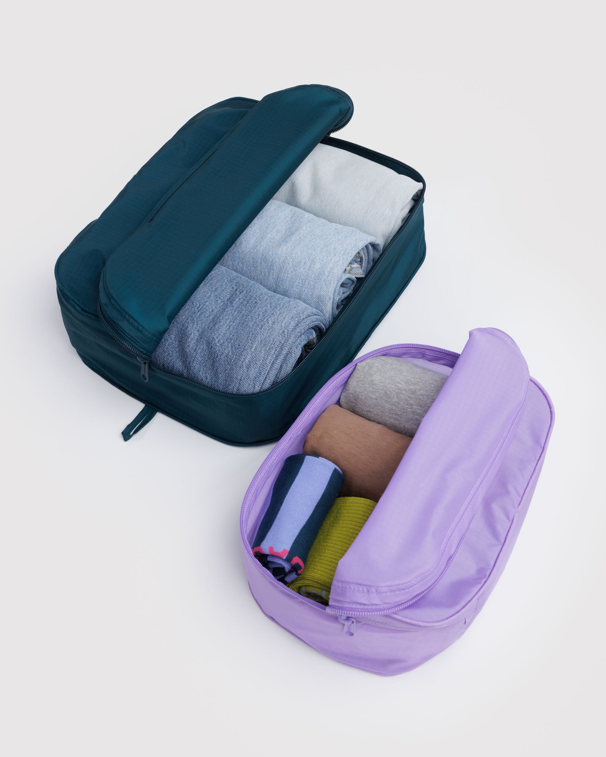 Two travel packing cubes, one teal and one purple, filled with clothes and zip top half folded back.