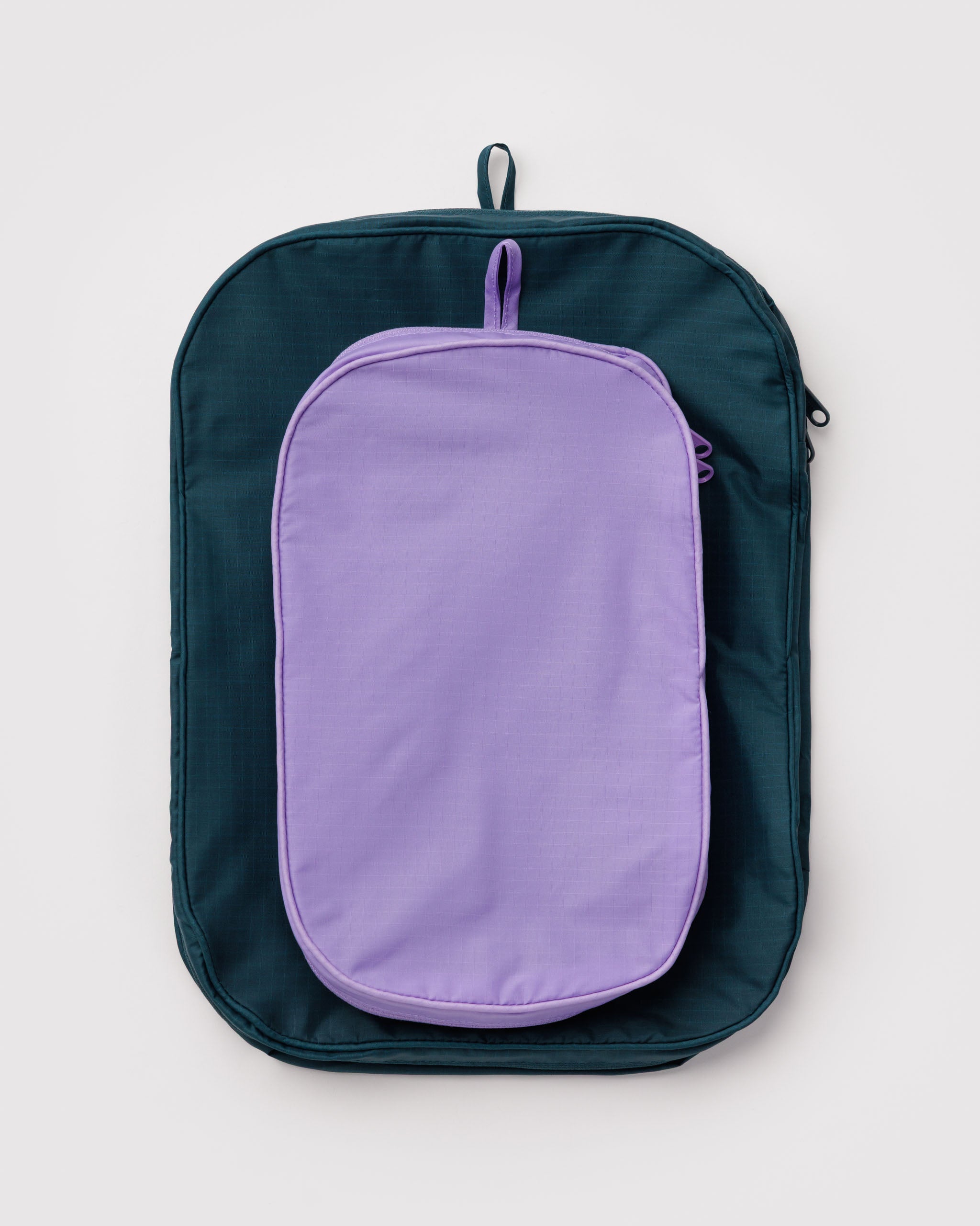 A small size purple and medium size teal packing cube collapsed flat for storage with hanging loop at the top and double zips closed.