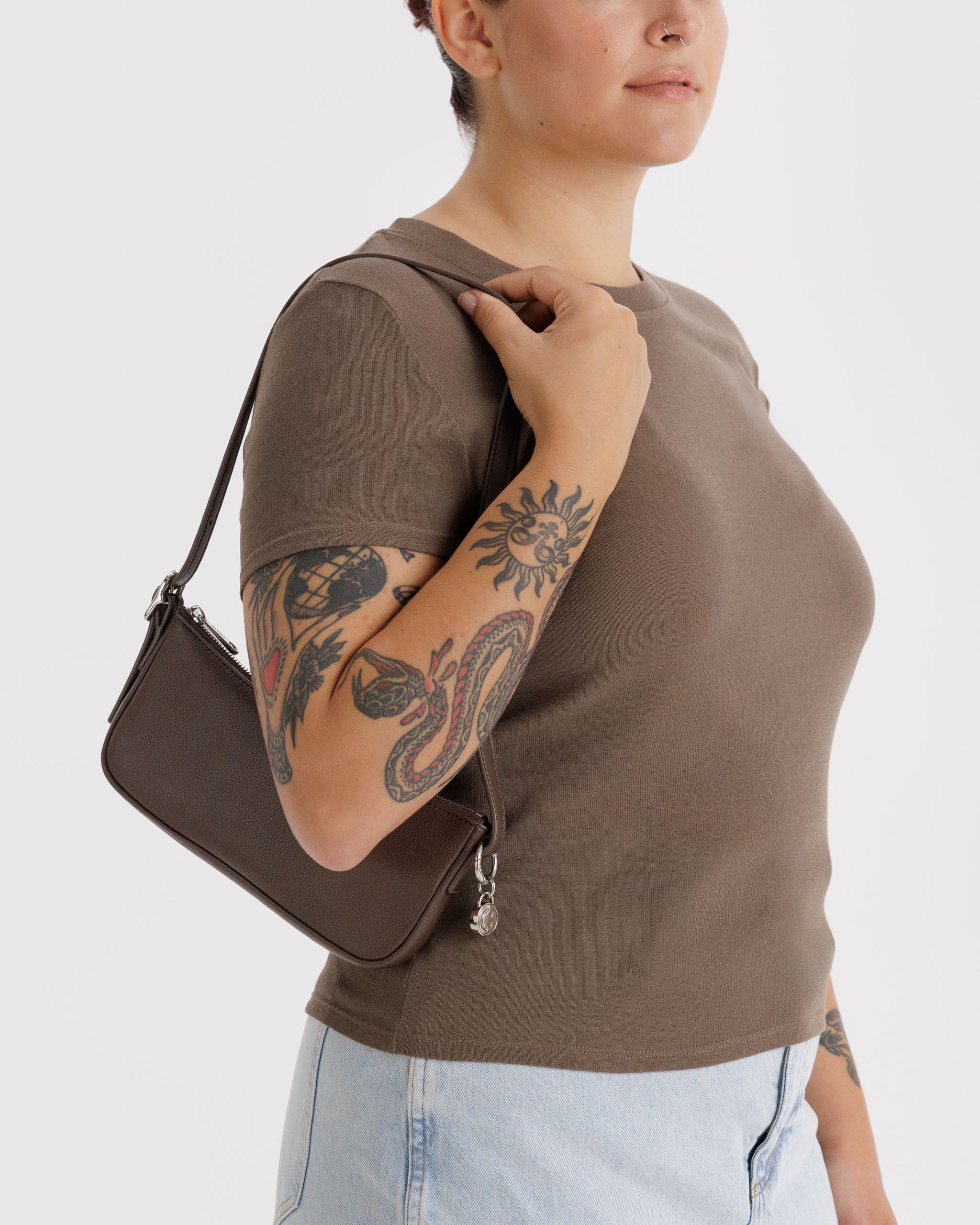 Person wearing a brown shirt holding a matching brown leather bag with an attached metal silver smiley face charm, top zip and adjustable clasp for shoulder strap.