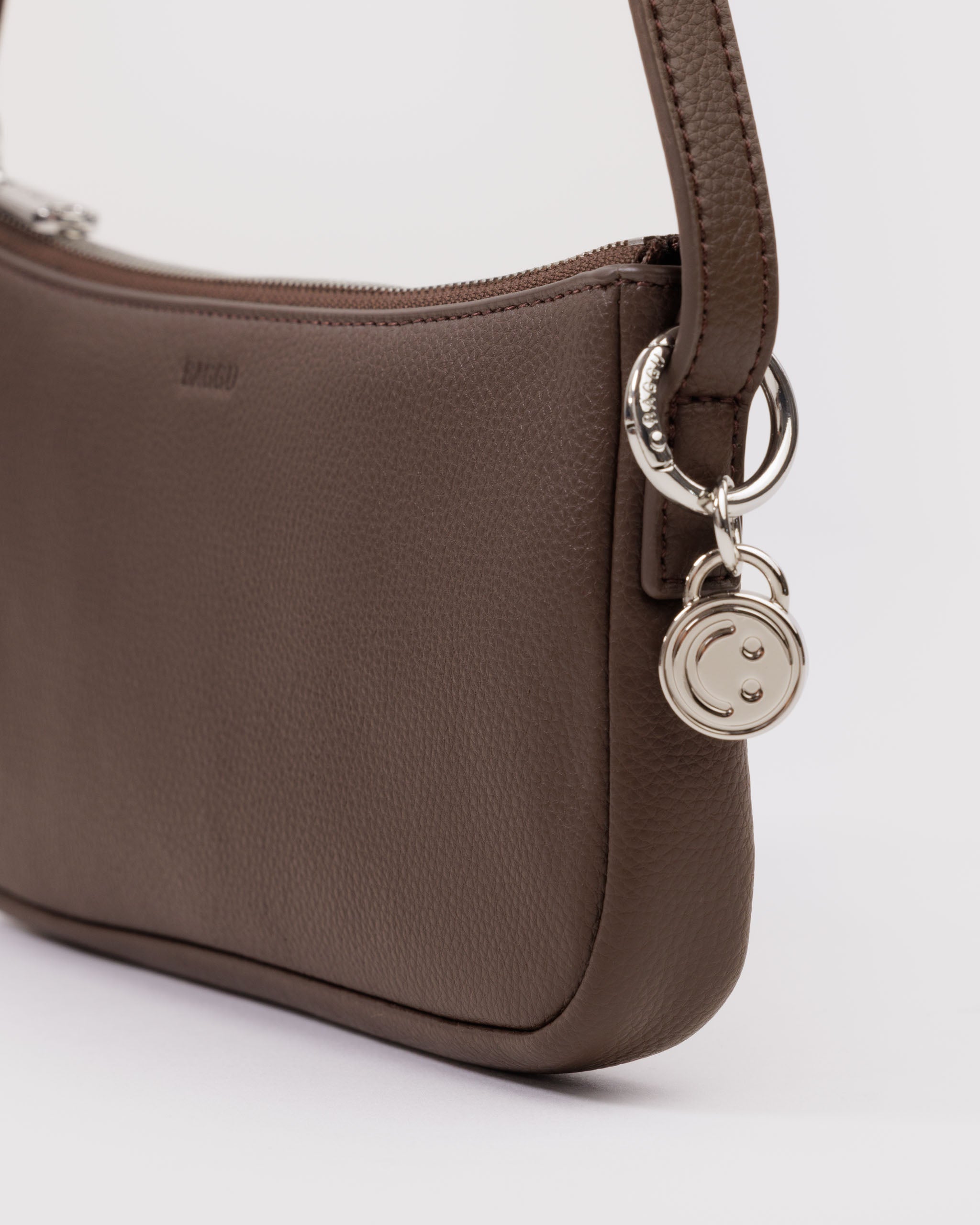 Brown leather handbag with a metal silver smiley face bag charm attached to shoulder strap by a BAGGU branded metal silver ring.