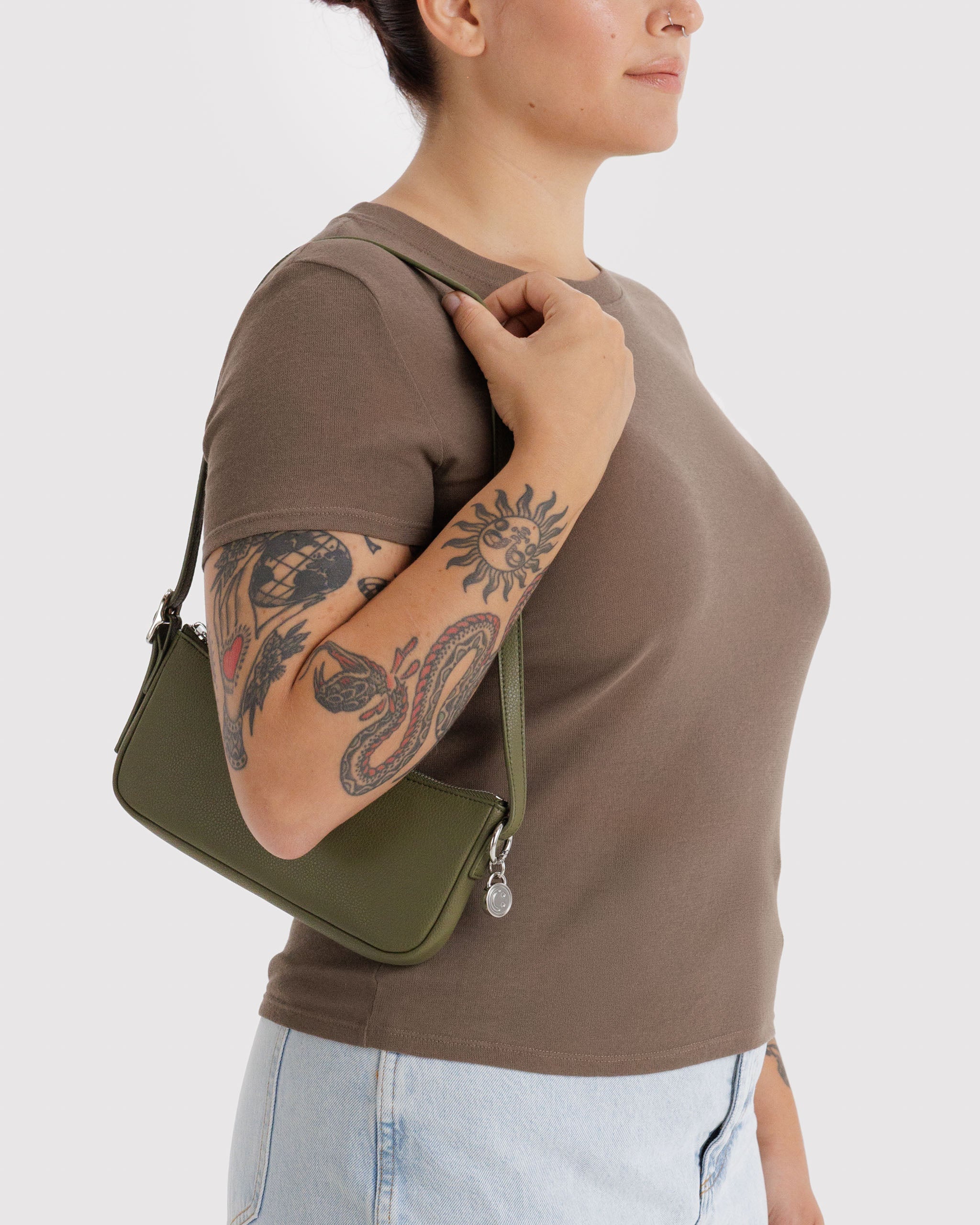 Person wearing a brown shirt and light blue jeans, holding a green leather handbag on shoulder with metal smiley face charm attached to adjustable strap.