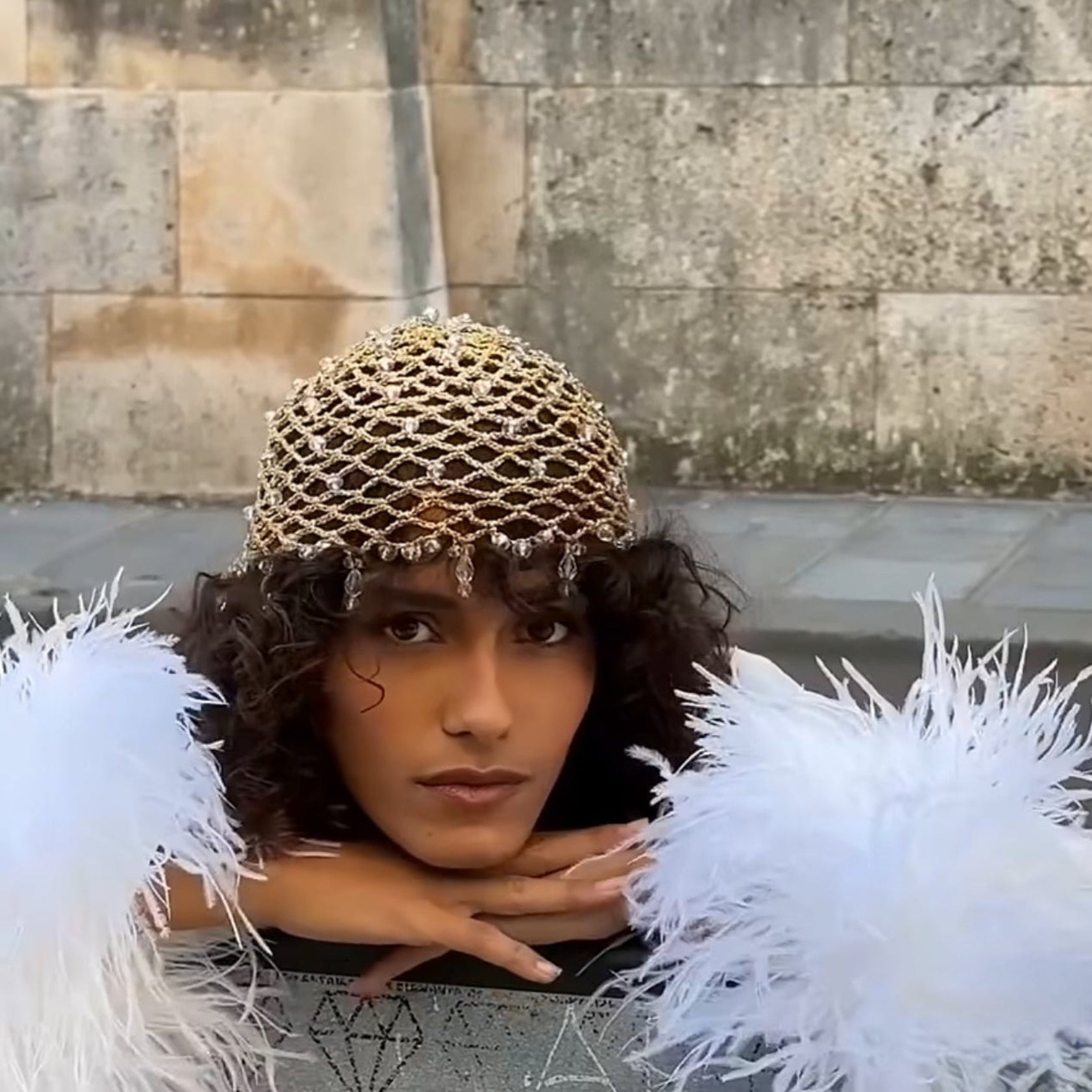 Person wearing a gold glass crystal beaded hat and white feathers against a stone wall.