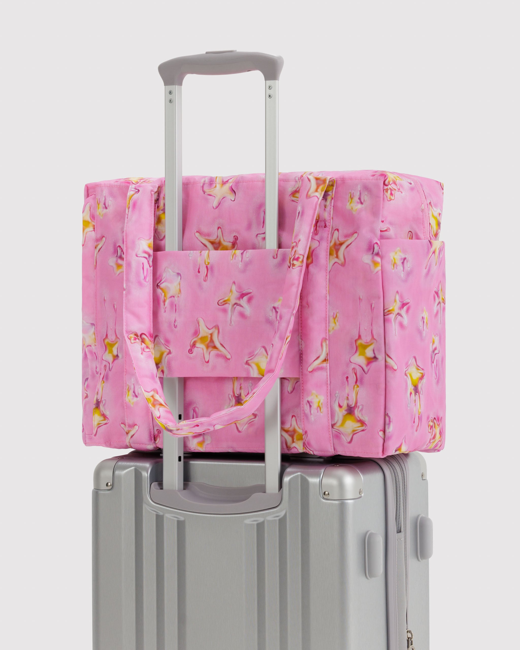 Pink zip tote bag with yellow melting stars pattern on top of a silver suitcase attached with trolley sleeve.