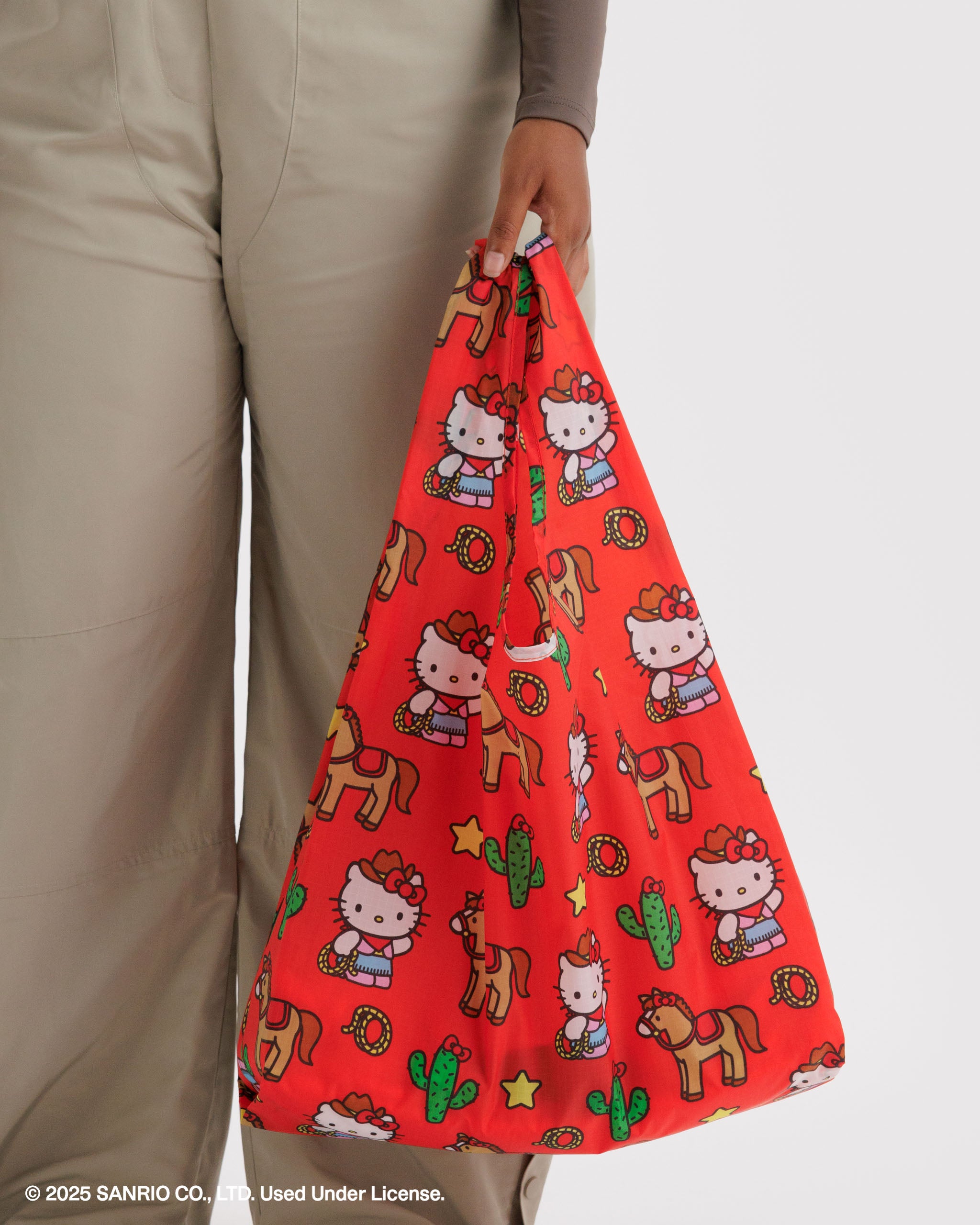 Red nylon tote bag with Hello Kitty and horse pattern held by a person wearing beige pants.
