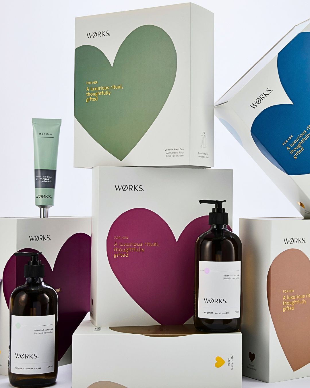 A stack of Works skincare product boxes with heart-shaped designs and writing surrounded by glass bottles with pumps and a green tube.